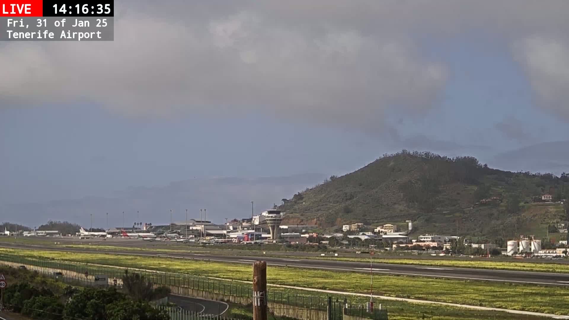A long shot of an airport runway with several planes, surrounded by low-lying vegetation and a hill in the background, under an overcast sky.