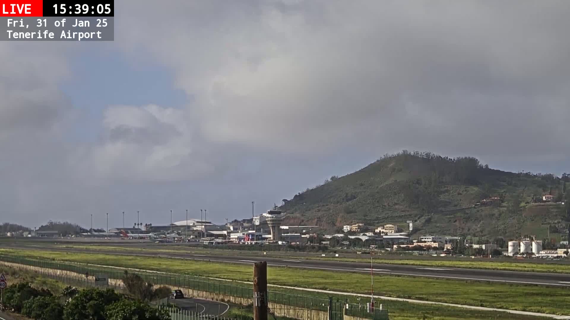 An airport runway and terminal buildings are visible beneath an overcast sky, with a large green hill in the background.