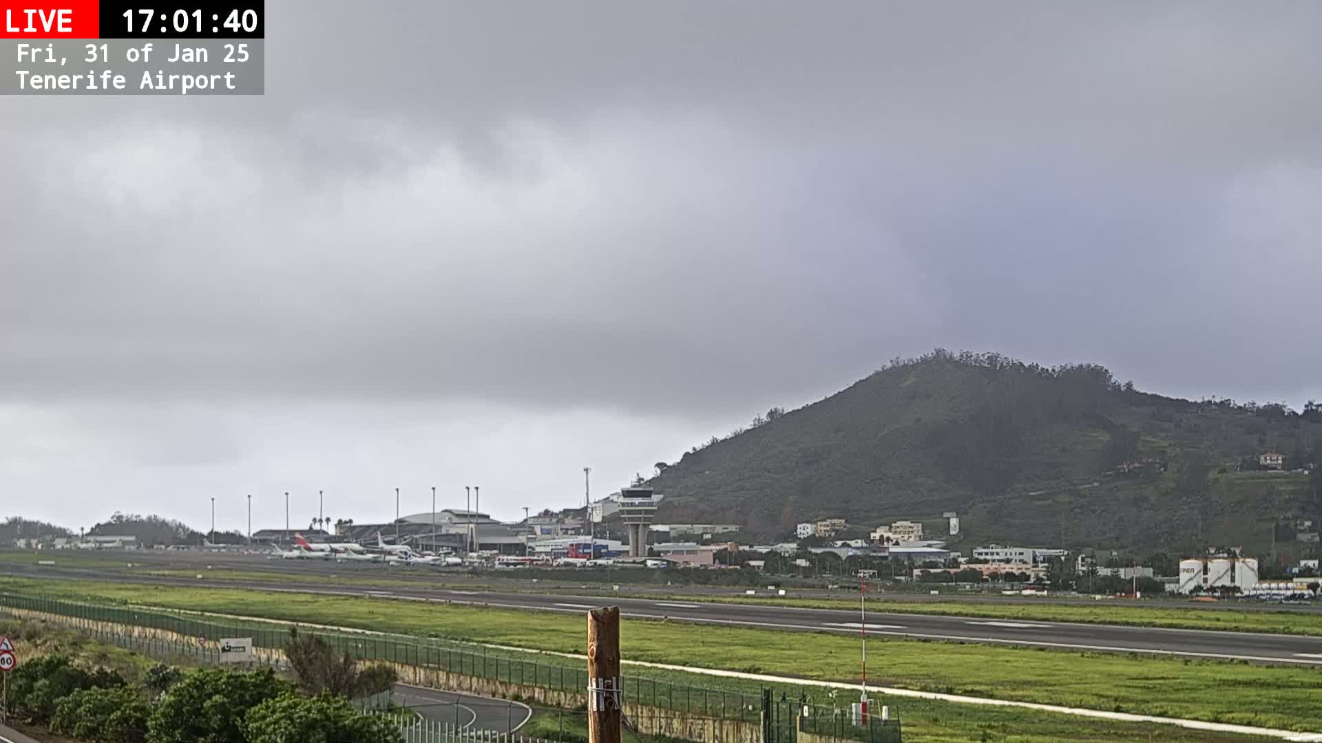 A view of an airport runway with several airplanes parked, a control tower, and a green hill in the background under an overcast sky.
