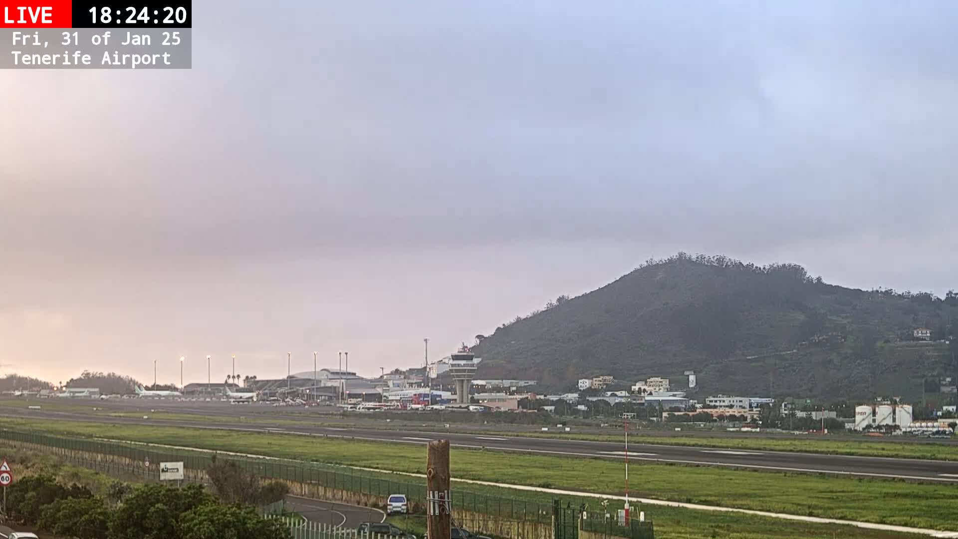 A view of an airport runway with several planes on the tarmac, under a hazy, overcast sky, with a hill visible in the background.