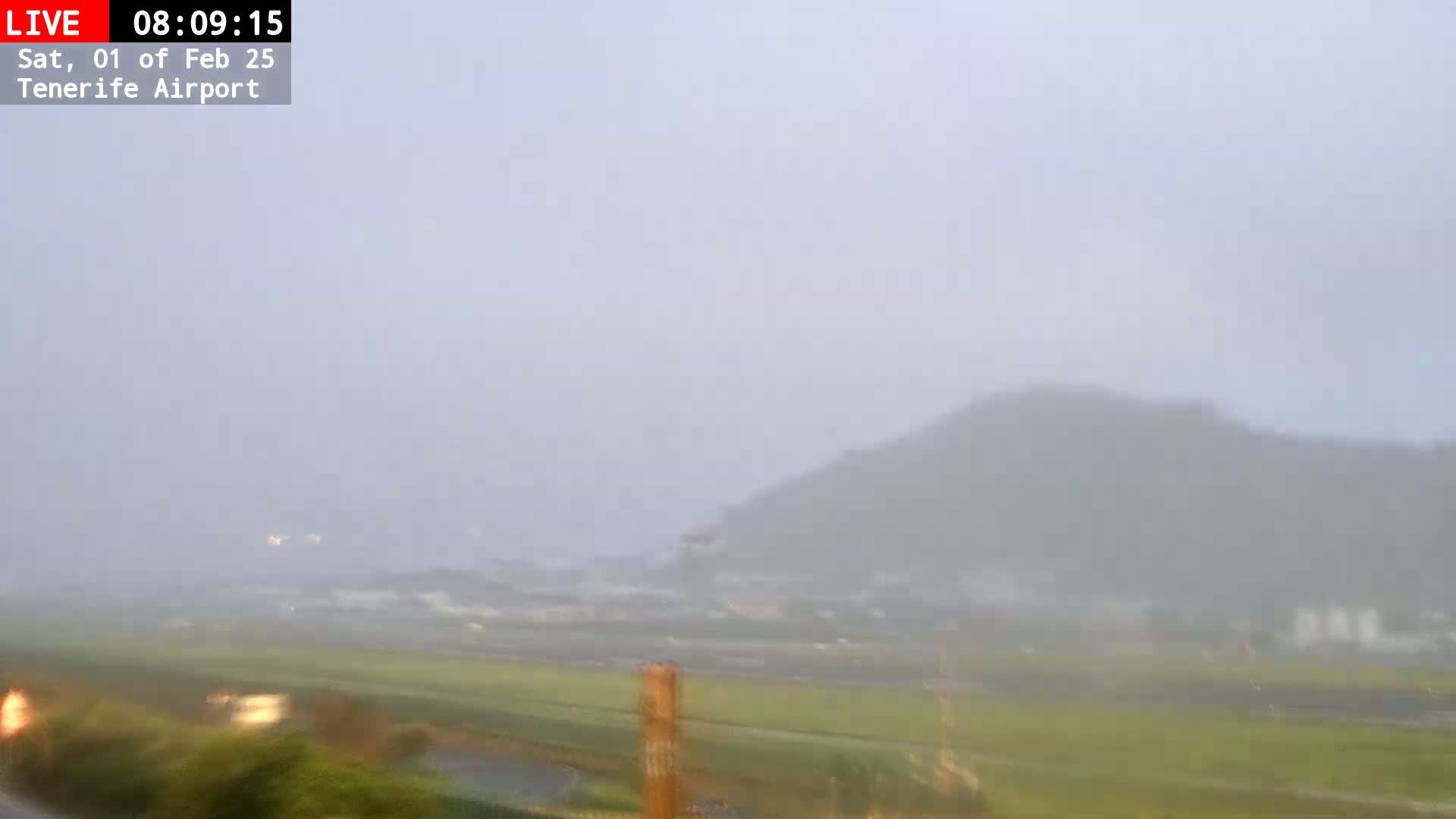 A hazy, rainy view shows a distant hill and flatlands with some structures in the foreground.