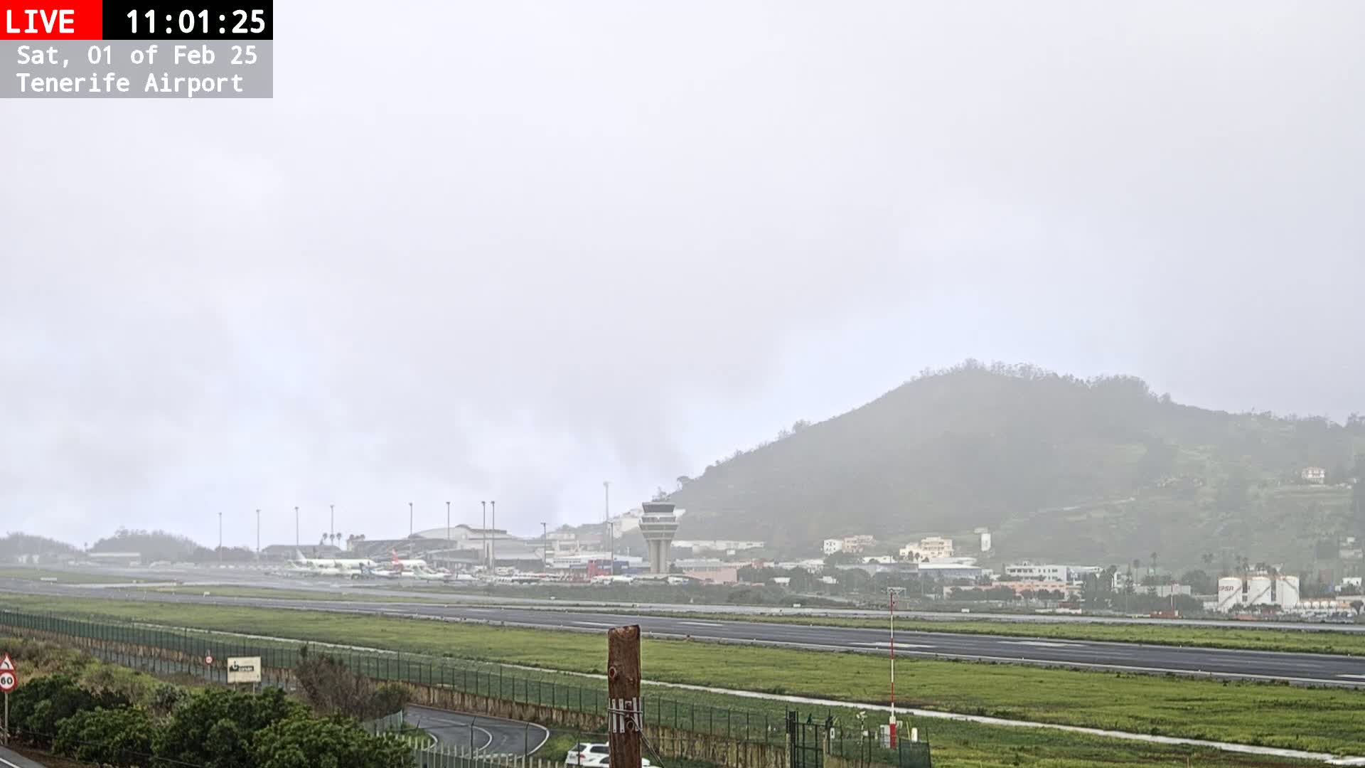 A foggy view of an airport runway, control tower, and airplanes, with a hill in the background.