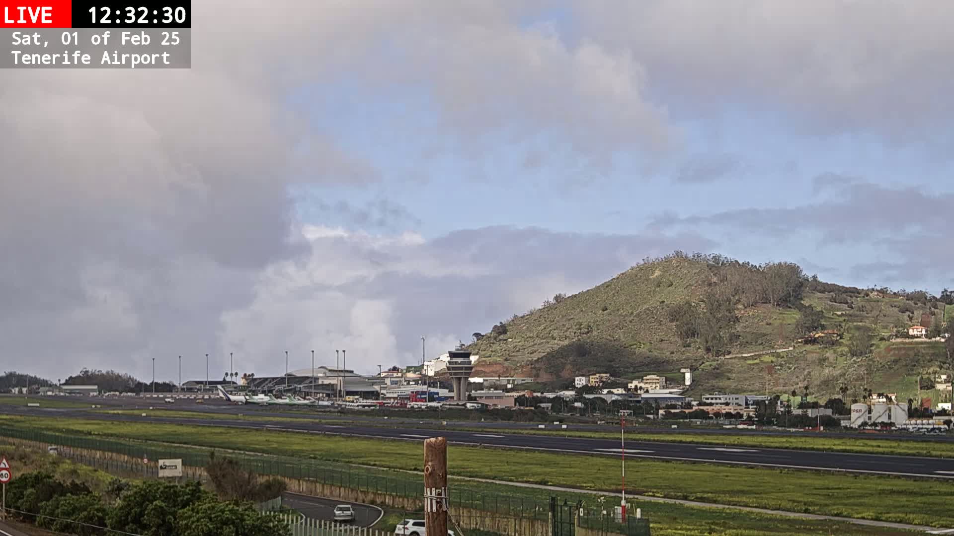 A mostly cloudy sky overlooks an airport with several planes on the tarmac, a control tower, and a green hill in the background.