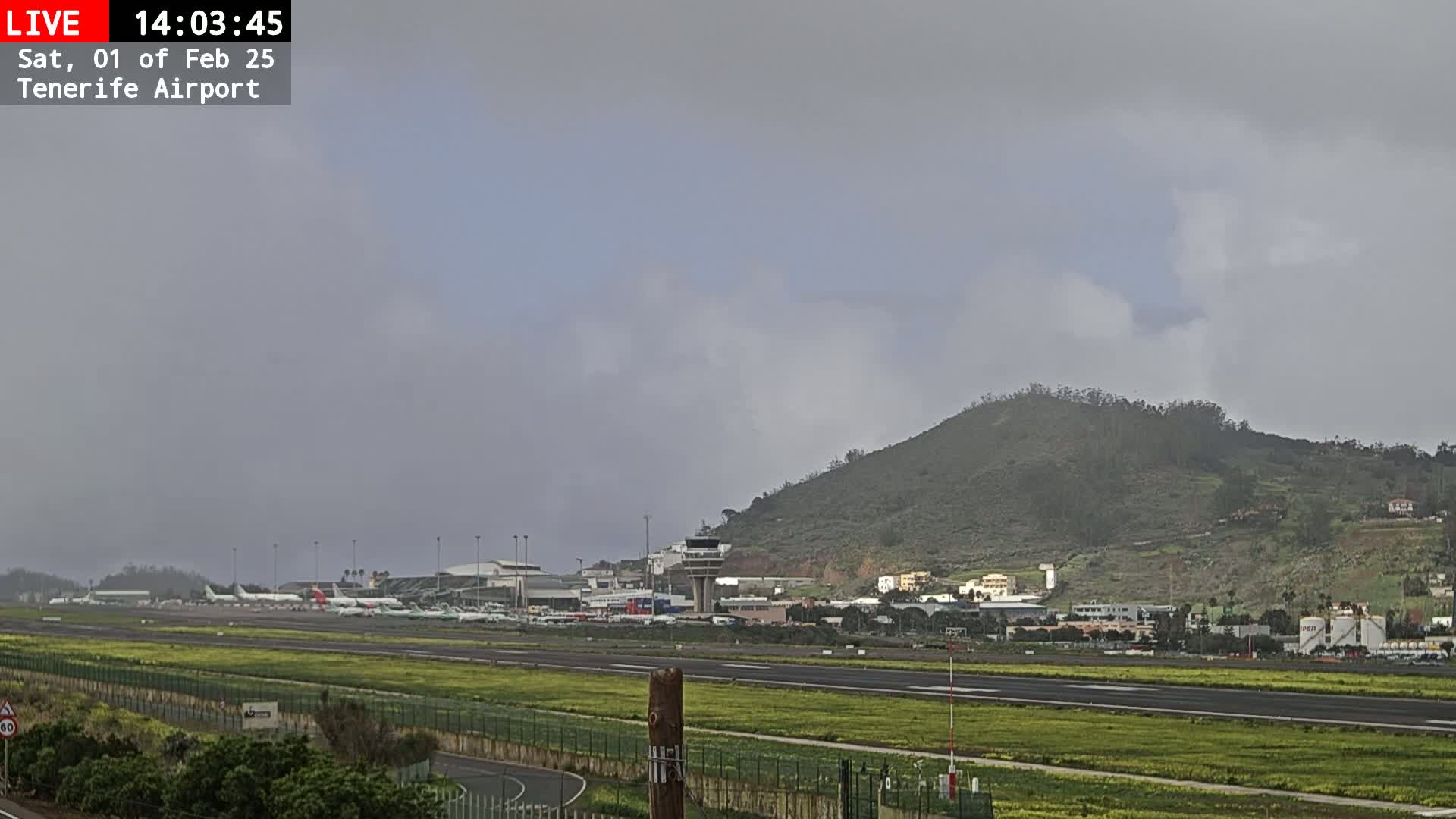 A mostly cloudy sky overlooks an airport with numerous airplanes parked on the tarmac, situated next to a green hill.