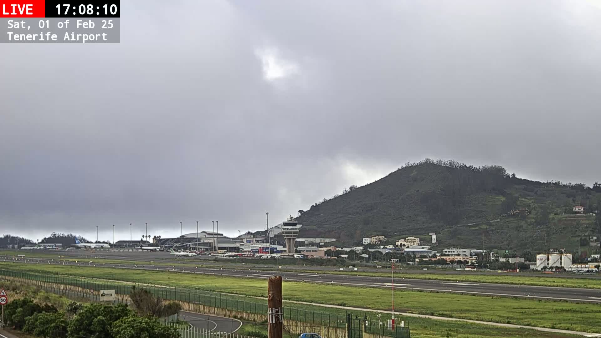 A view of an airport runway and terminal buildings under an overcast sky, with a hill in the background.