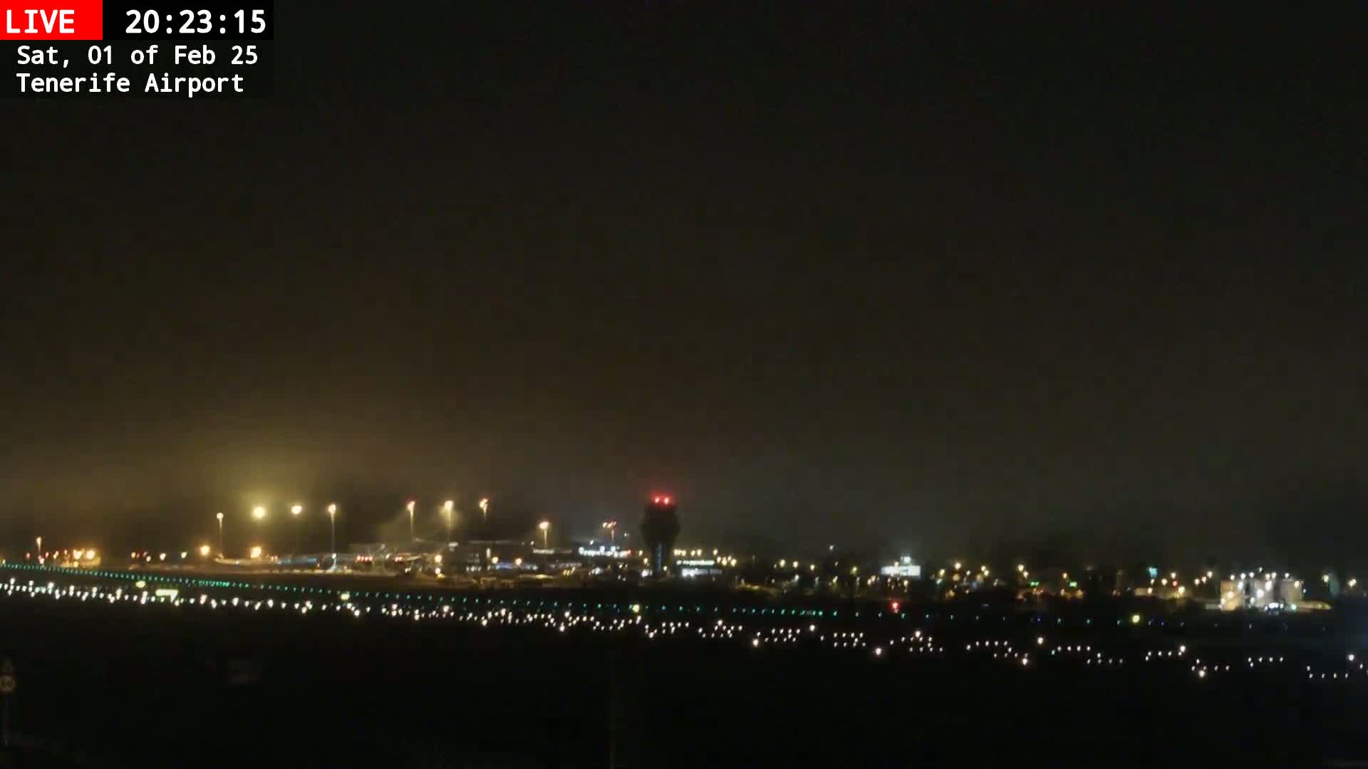 A nighttime view of an airport runway and terminal buildings under foggy conditions.