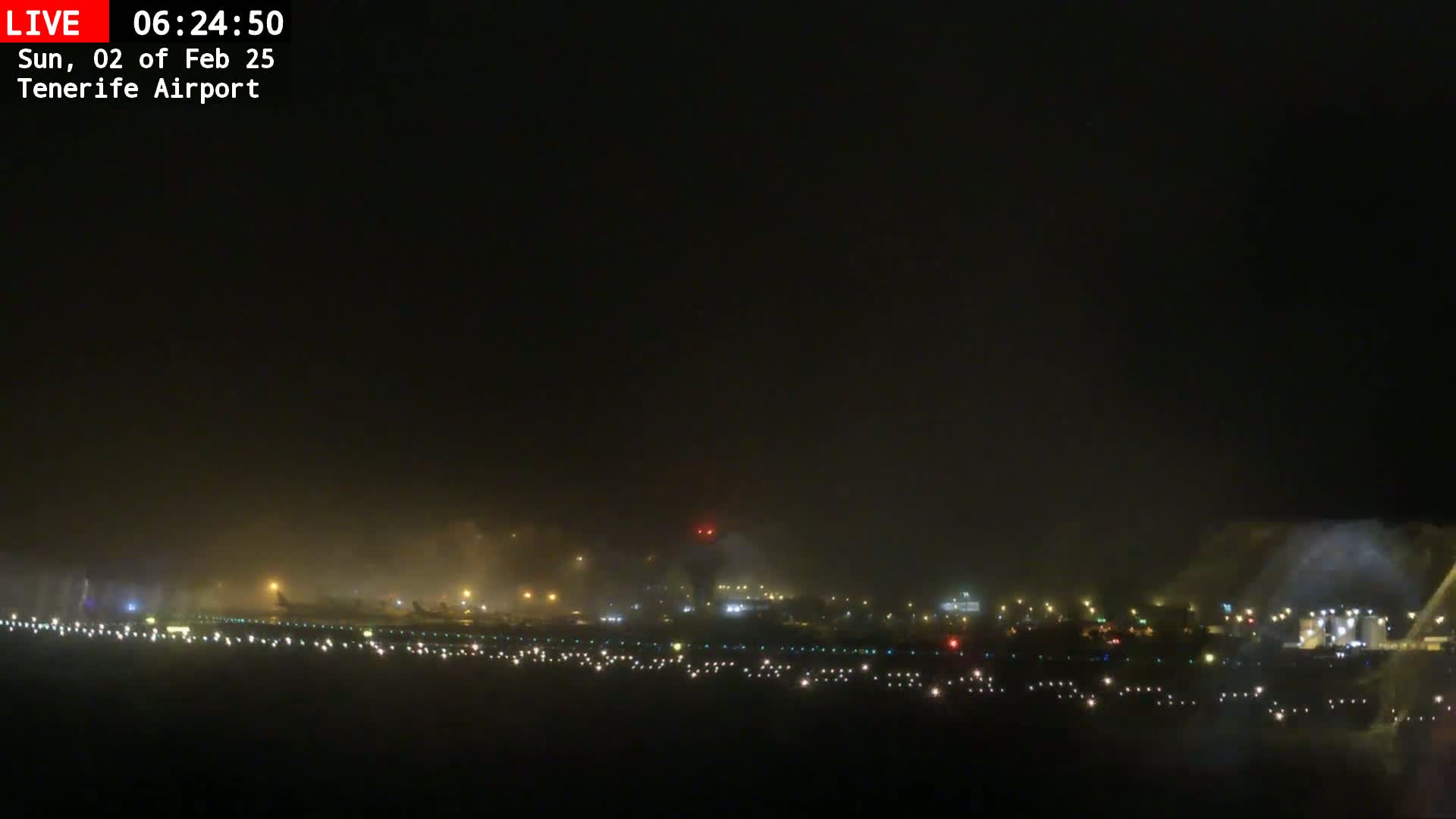 A foggy nighttime view of an airport runway with dimly lit buildings and aircraft in the distance.