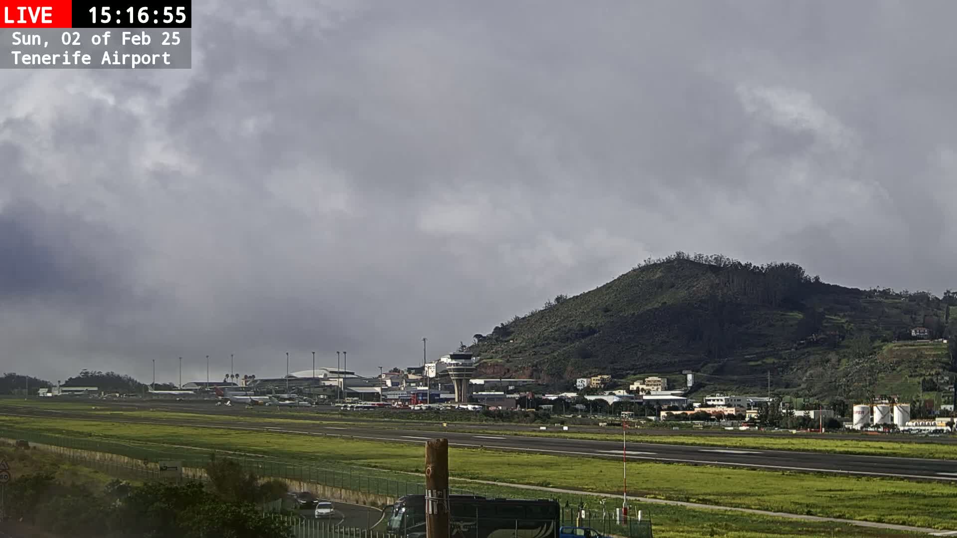 A view of an airport runway with several planes visible, a control tower, and a large hill in the background under an overcast sky.
