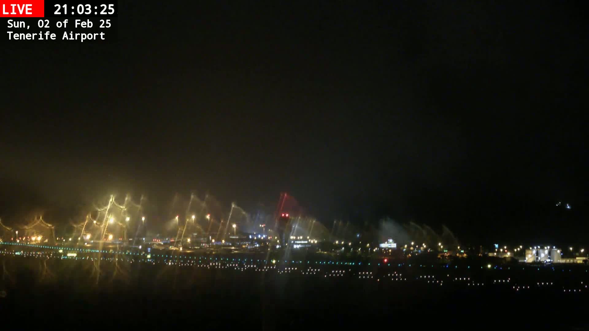 A nighttime view of an airport runway with lights illuminating water sprays, under a dark sky.