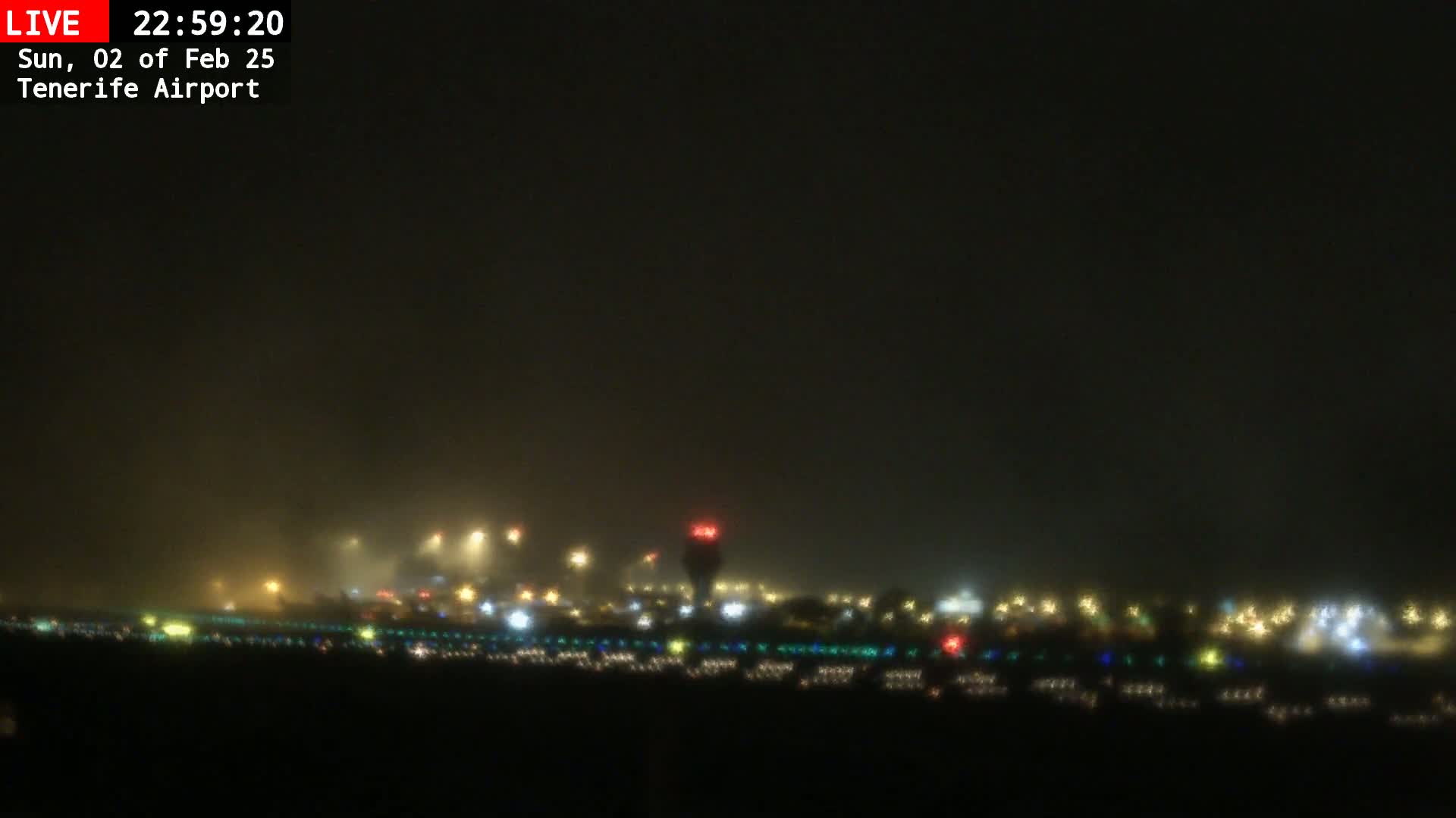 A hazy night scene shows a distant airport runway and buildings illuminated with various colored lights.