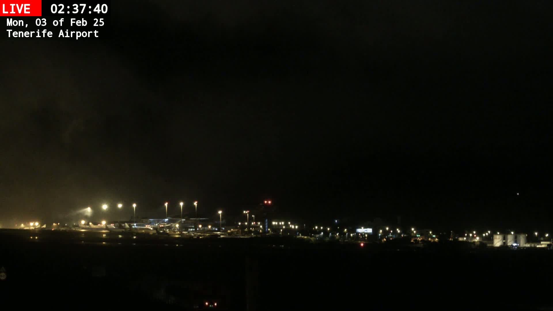 A nighttime view of an airport runway and terminal buildings under a dark, possibly cloudy sky.