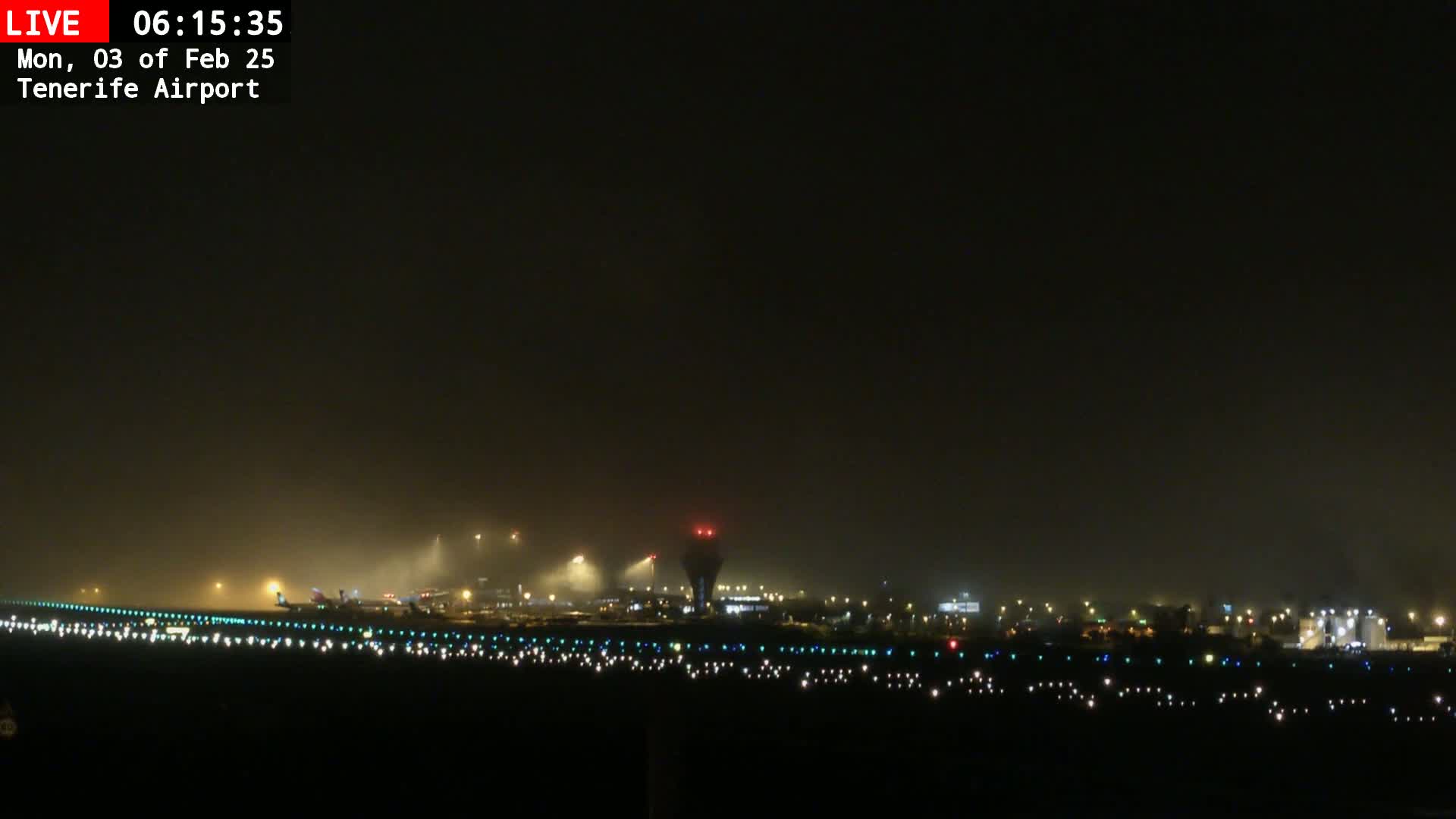 A foggy nighttime view of an airport runway with lights illuminating aircraft and the control tower.