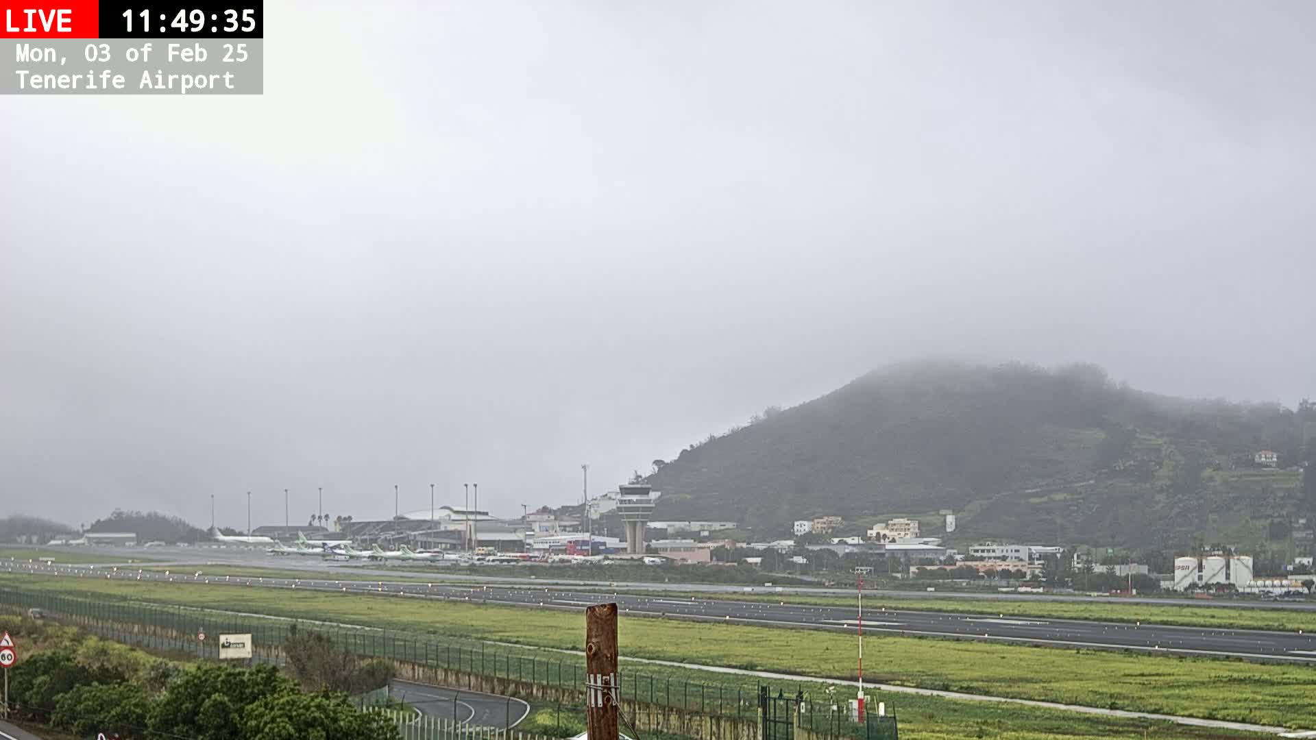 A foggy view of an airport runway with several airplanes parked, surrounded by grassy fields and a hill shrouded in mist.