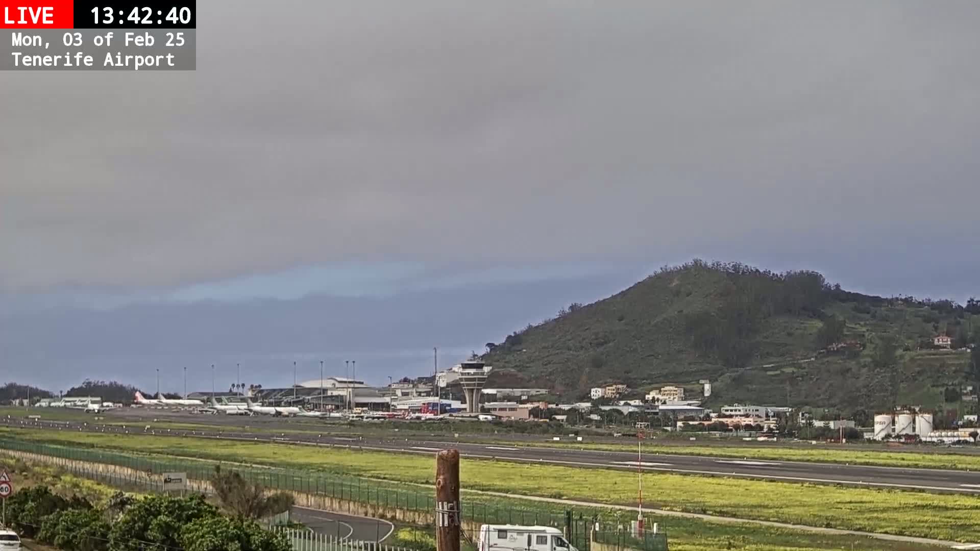 An airport runway with several airplanes, surrounded by low-lying hills and fields of yellow flowers under an overcast sky.