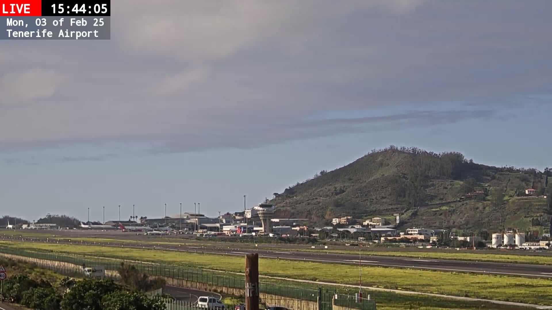 A mostly cloudy sky overlooks an airport runway with planes and buildings, beyond which is a large, sparsely vegetated hill.