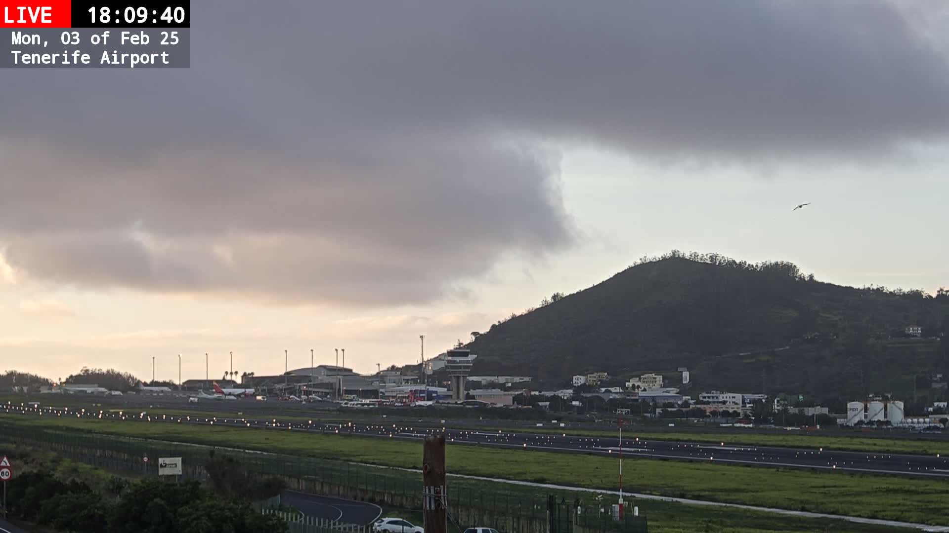 An overcast sky hangs over an airport runway and control tower, with a hill rising in the background.