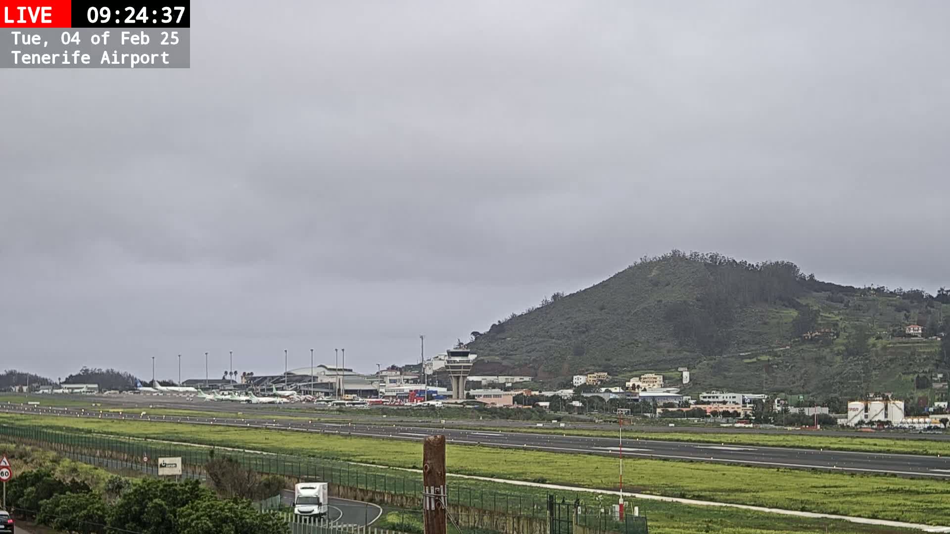 A view of an airport runway with several airplanes parked, under an overcast sky, with a green hill in the background.