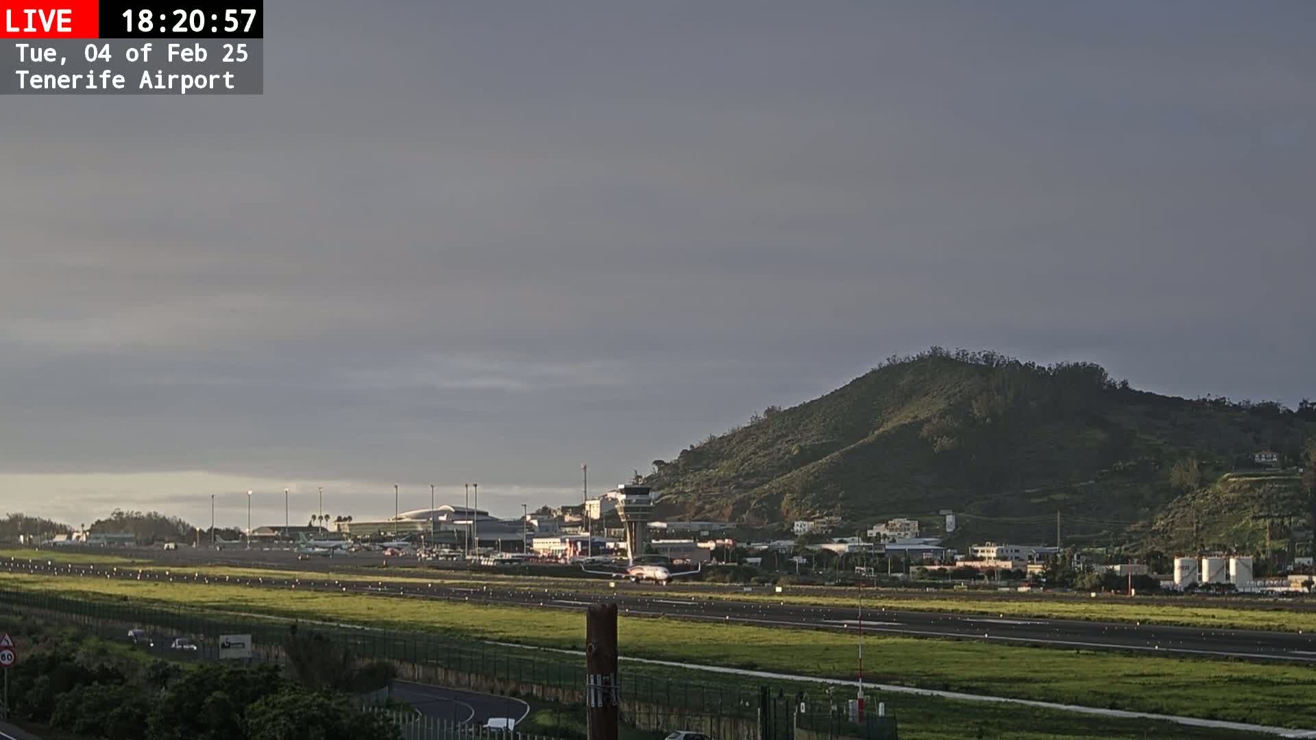 An airplane taxis on a runway at an airport near a large hill under a mostly cloudy, dusky sky.