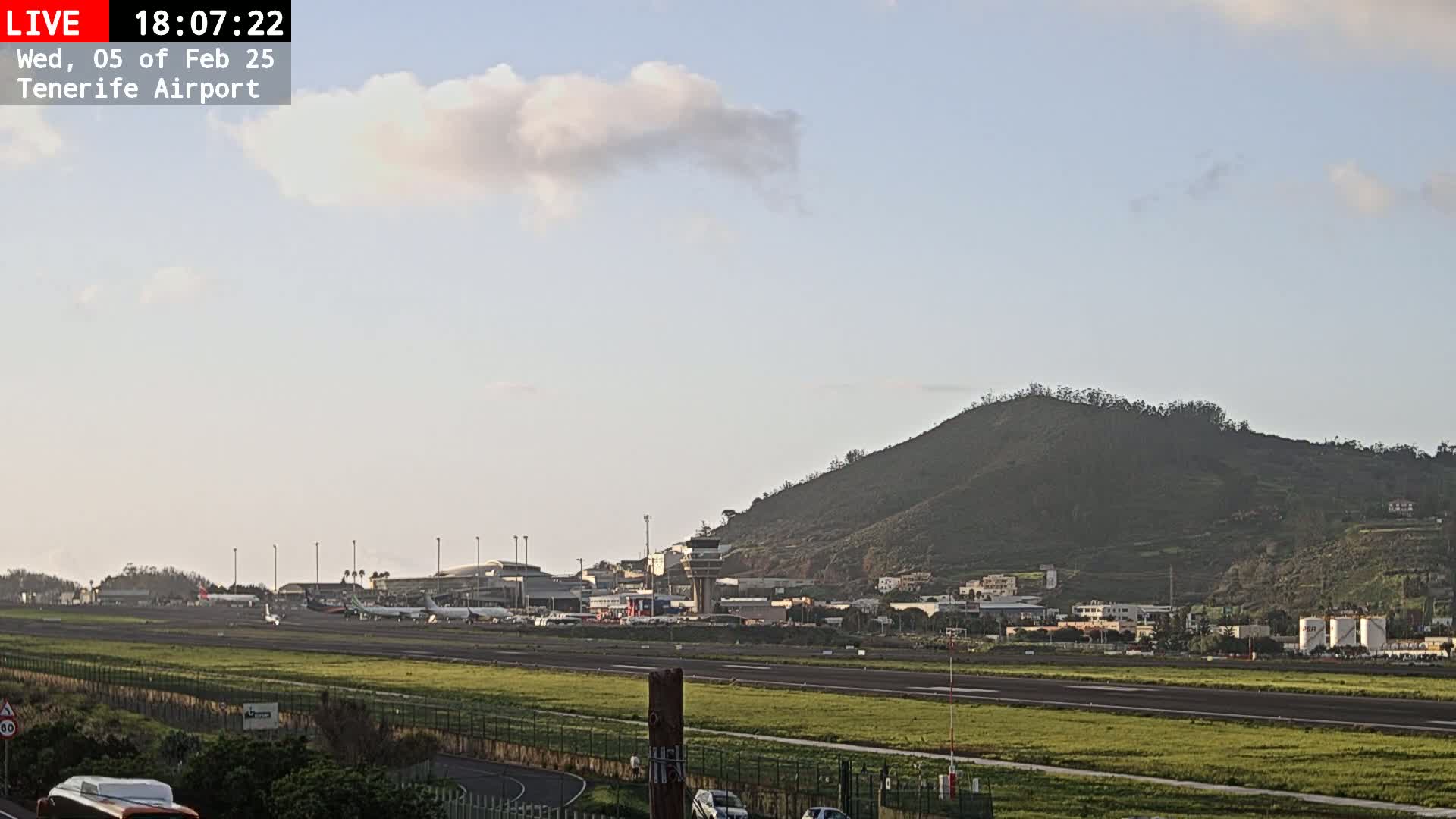 A mostly clear sky with a few clouds hangs over an airport with several airplanes on the tarmac, next to a grassy field and a large hill.