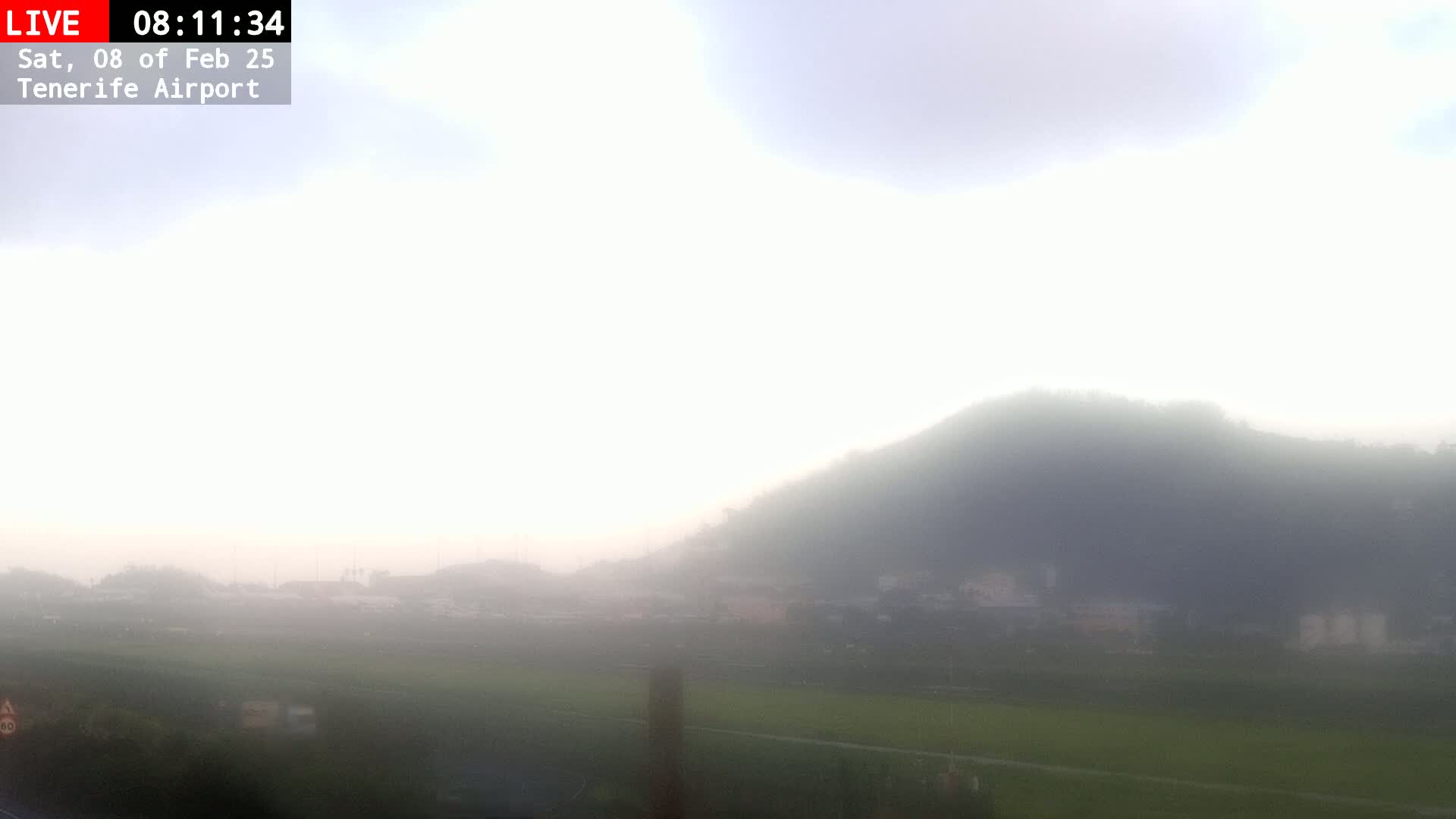 A foggy landscape reveals a hazy hill overlooking a flat expanse of green field and distant buildings.