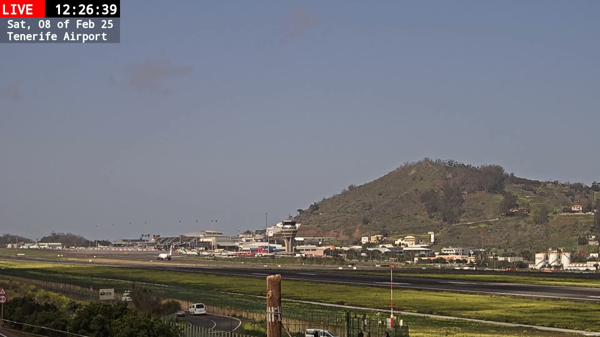 An airport runway and terminal are visible beneath a clear, light blue sky, with a large hill in the background.