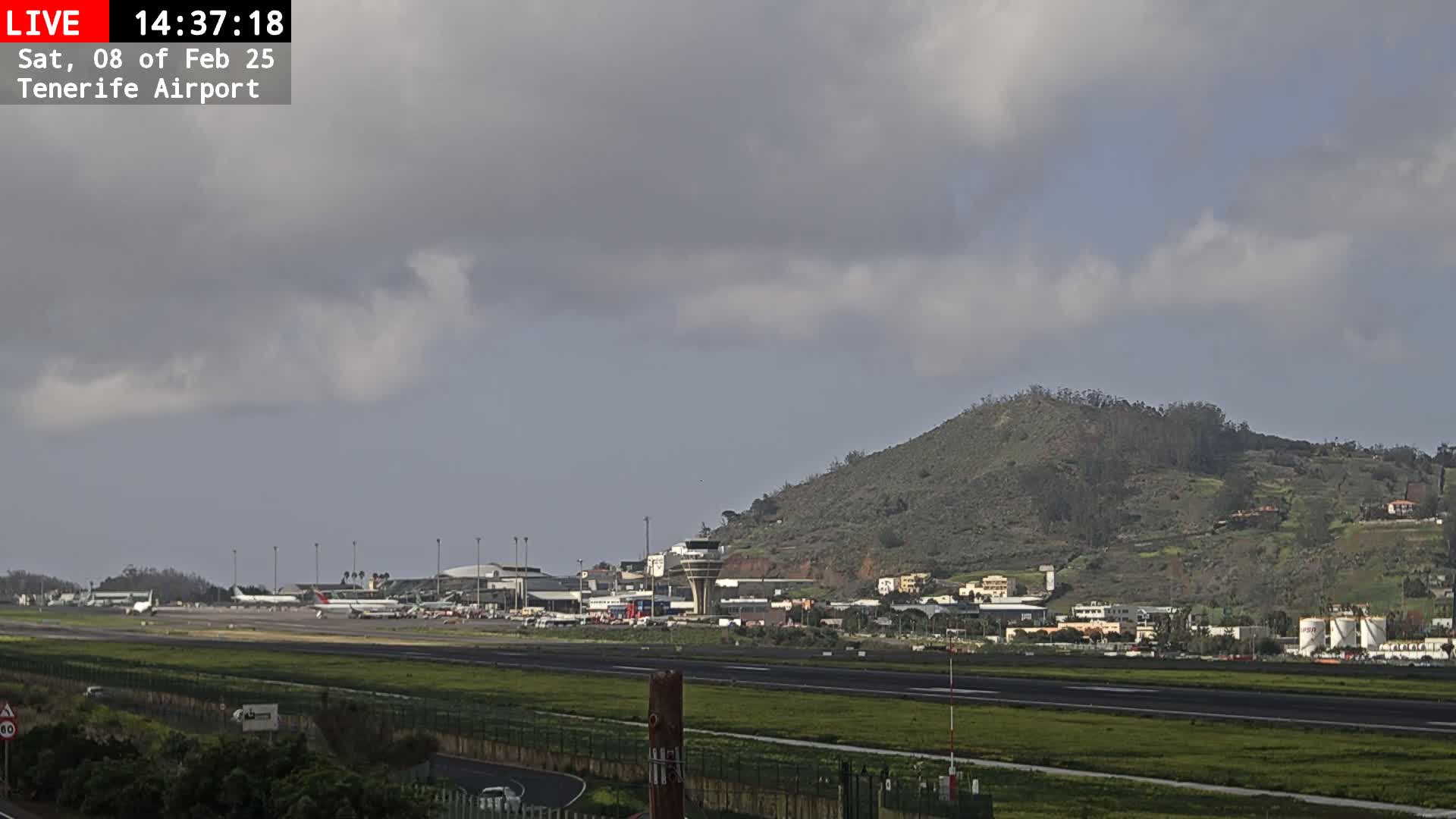 A mostly cloudy day at an airport with several planes on the tarmac, buildings, and a green hill in the background.