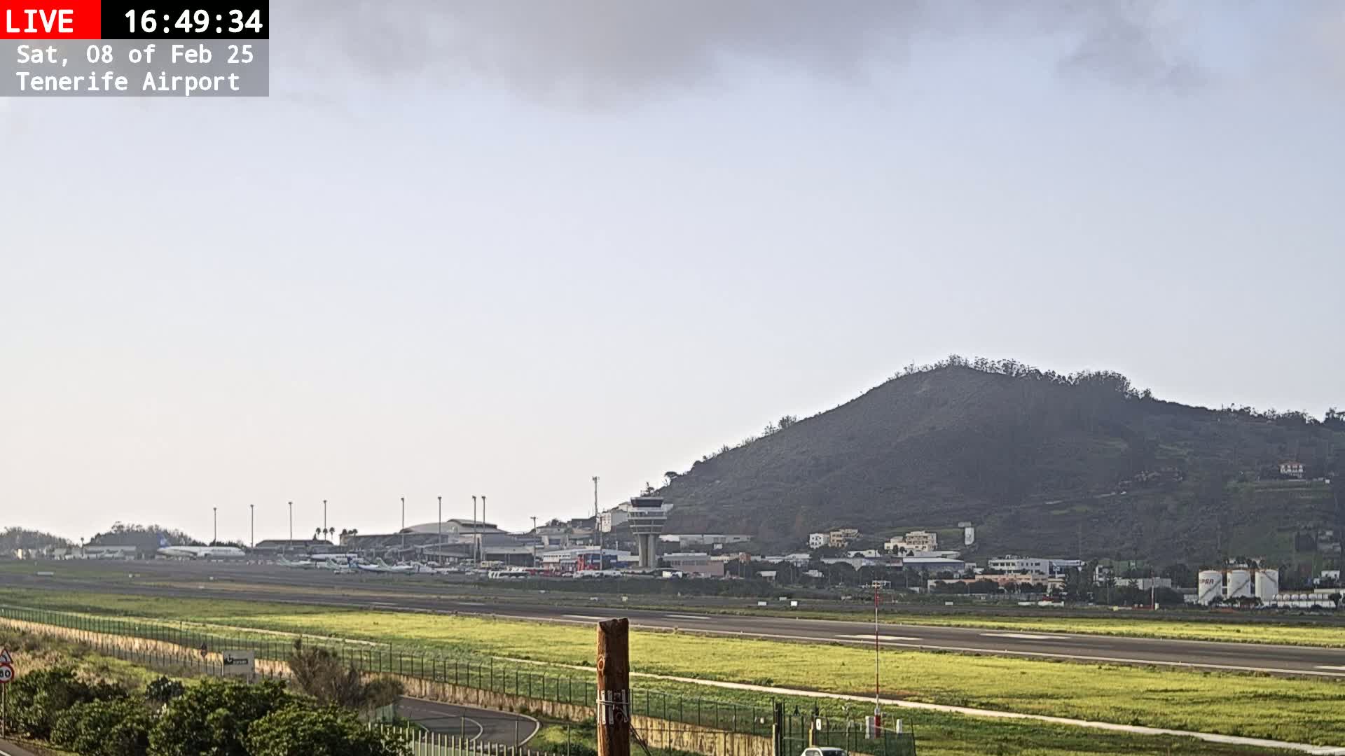 A long shot of an airport runway with several airplanes visible, a control tower, and a large hill in the background under an overcast sky.
