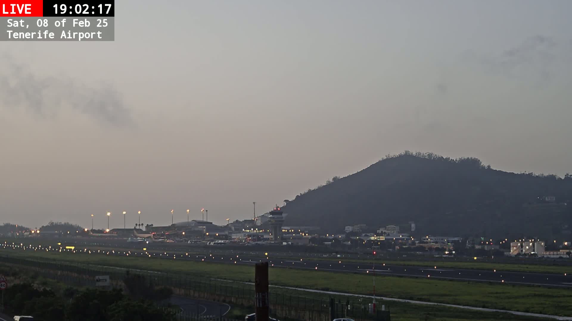 A long shot of an airport runway at dusk under a hazy sky, with a dark hill visible in the background.