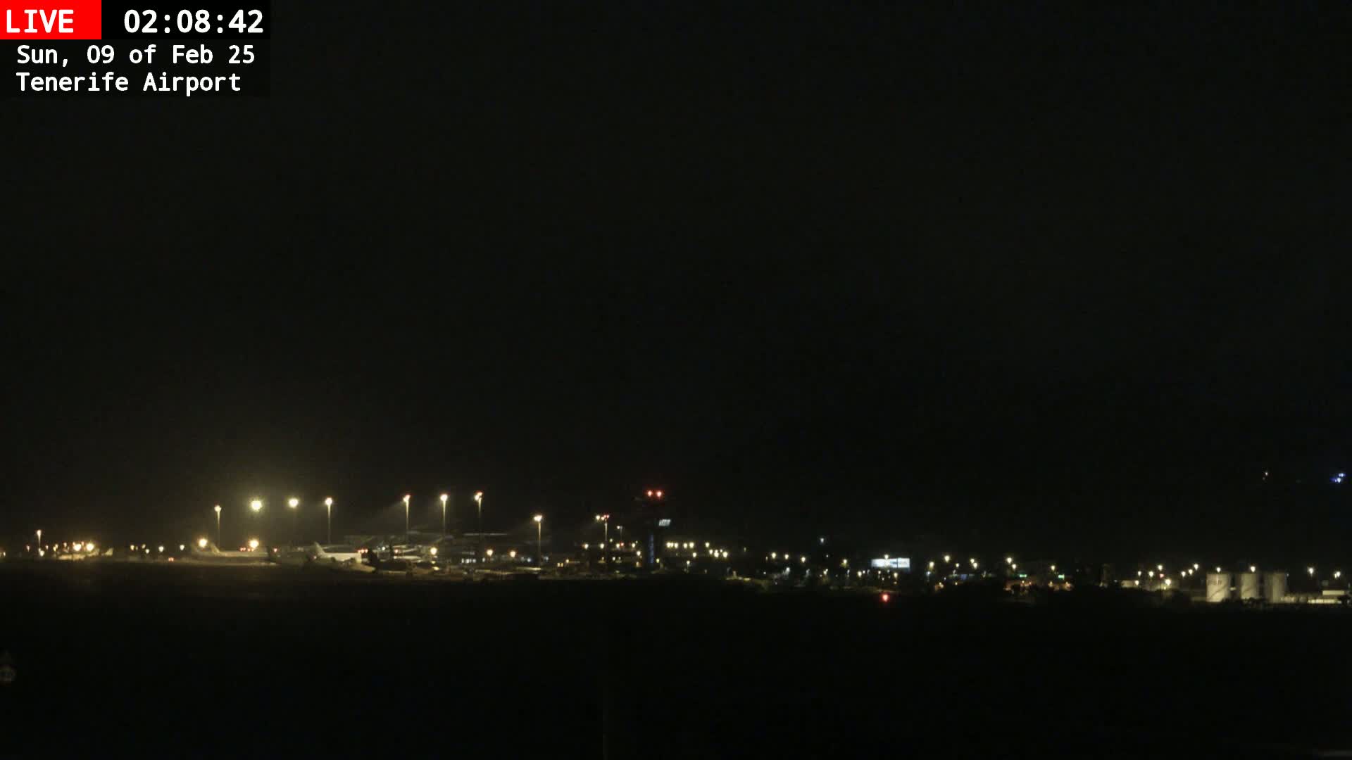 A nighttime view of an airport runway and terminal buildings illuminated by artificial lights under a dark, clear sky.