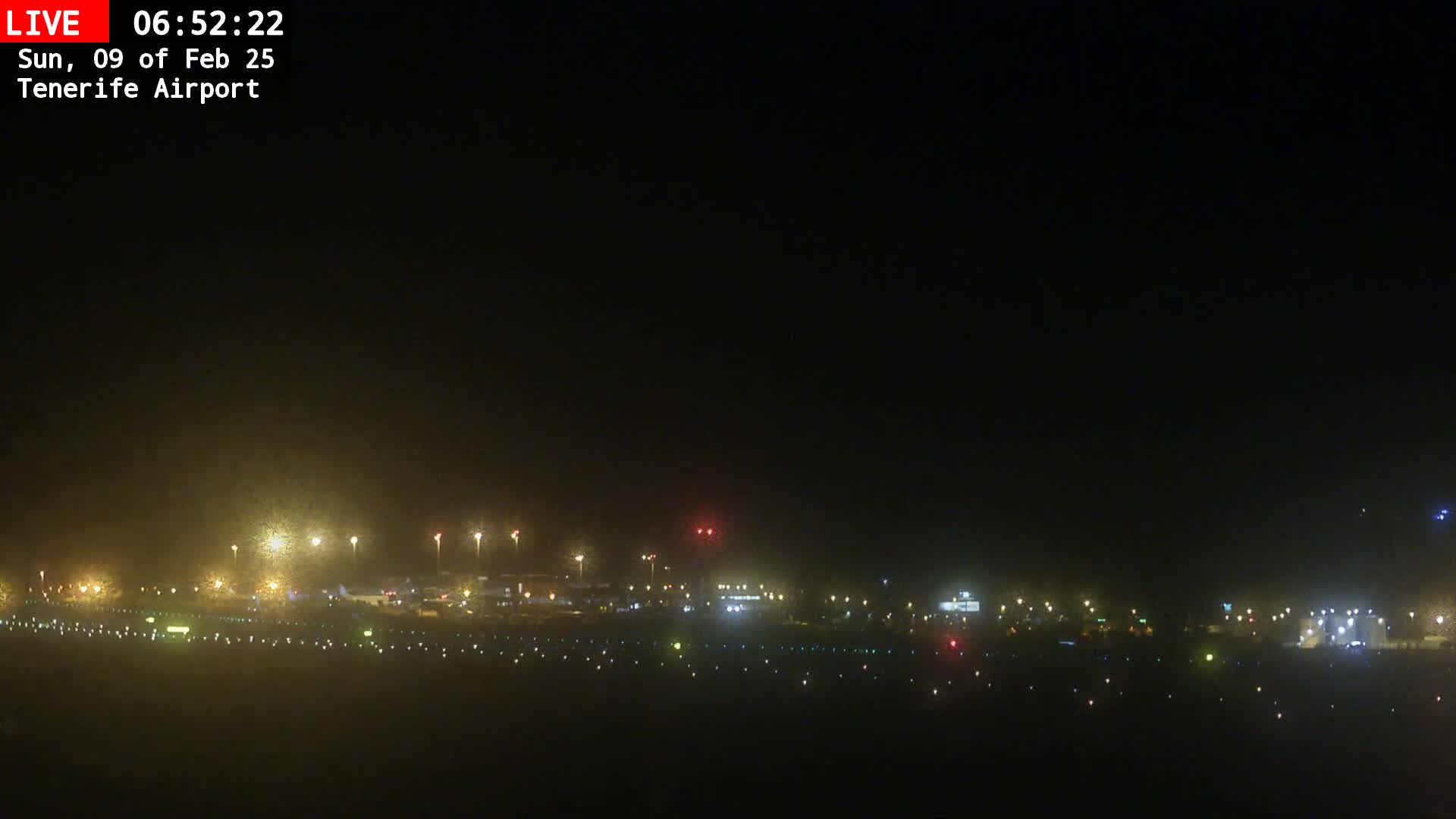 A nighttime long shot of an airport runway and tarmac, illuminated by numerous lights under hazy conditions.
