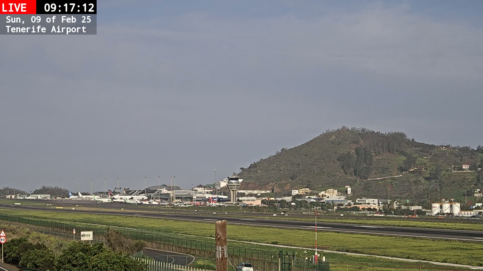 A mostly clear sky overlooks an airport with several planes parked on the tarmac, a control tower, and a grassy hill in the background.