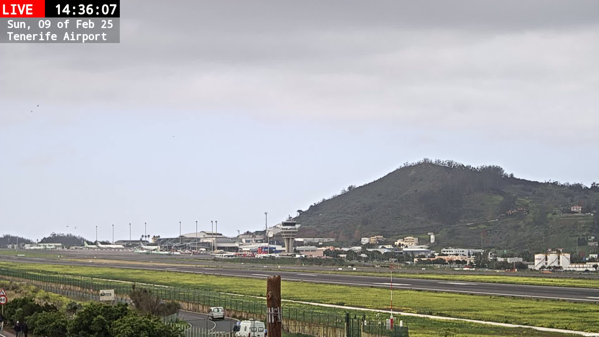 A long shot of an airport runway, control tower, and terminal buildings under an overcast sky, with a green hill in the background.