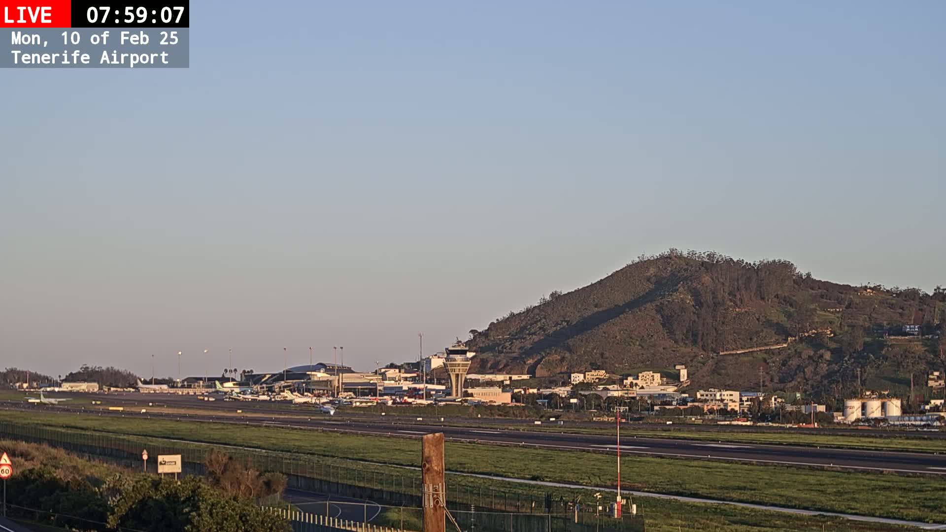 A long shot of an airport with several planes on the tarmac, under a clear, light-blue sky, with a hill in the background.
