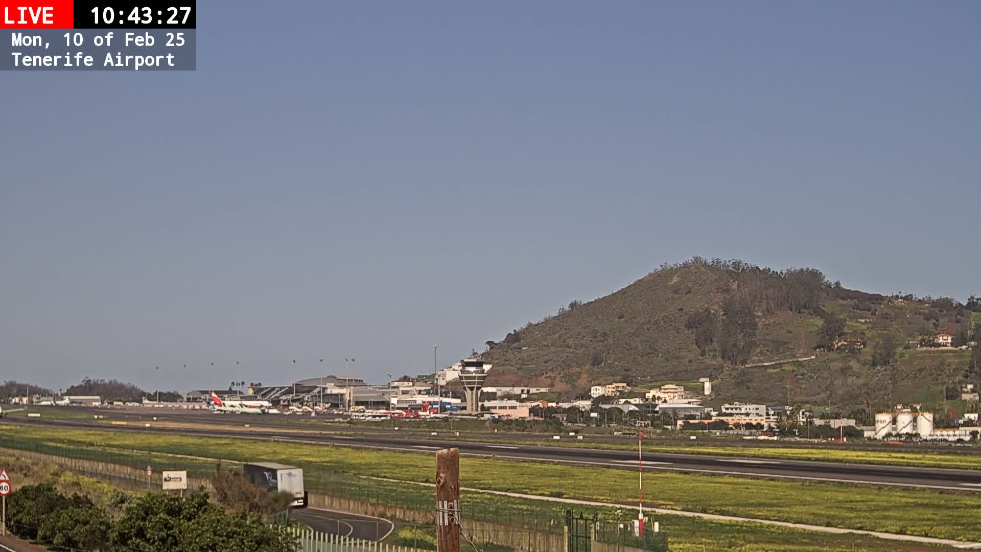 A sunny view of an airport runway with several airplanes parked near the terminal buildings, a hill in the background, and a truck driving on a road in the foreground.
