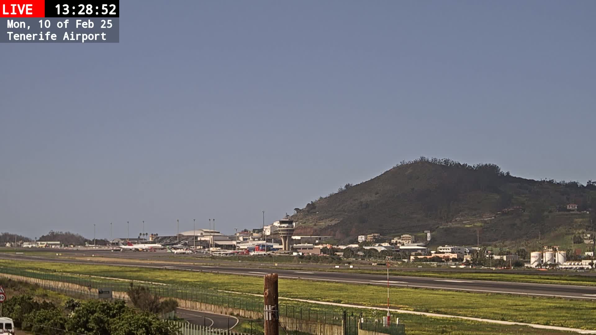 A sunny view of Tenerife Airport, featuring a runway, airport buildings, and a large hill in the background.