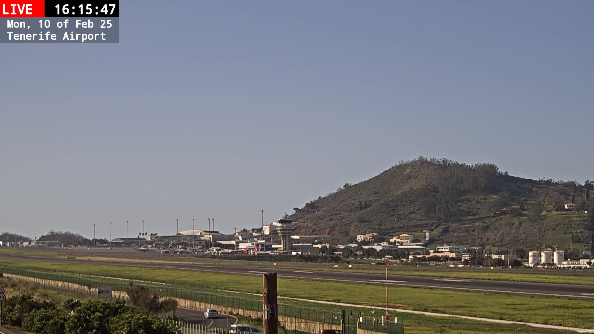 A long shot of an airport runway, control tower, and surrounding buildings under a clear sky, with a large hill in the background.