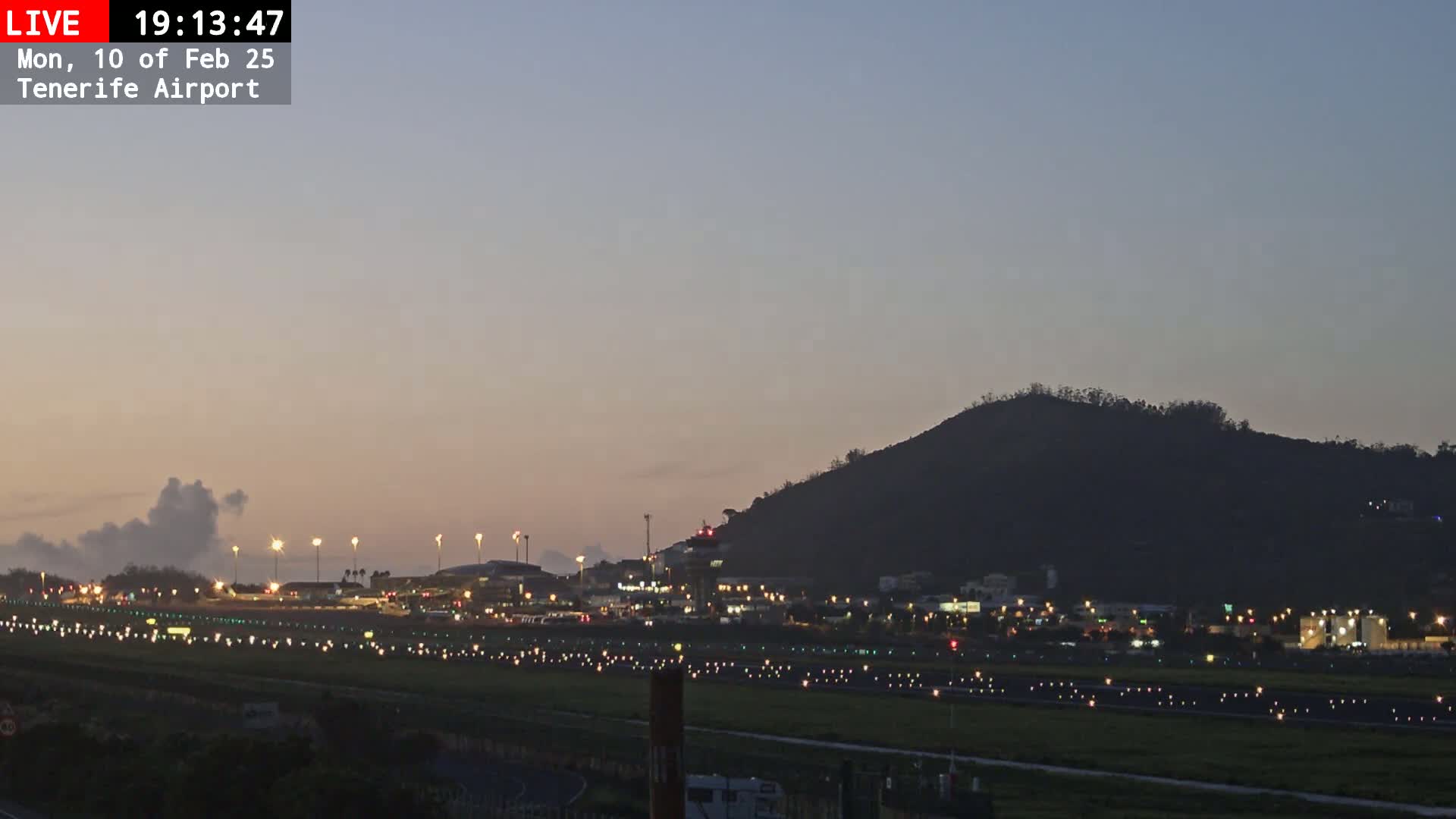A brightly lit airport runway at twilight, with a dark hill in the background under a mostly clear sky.