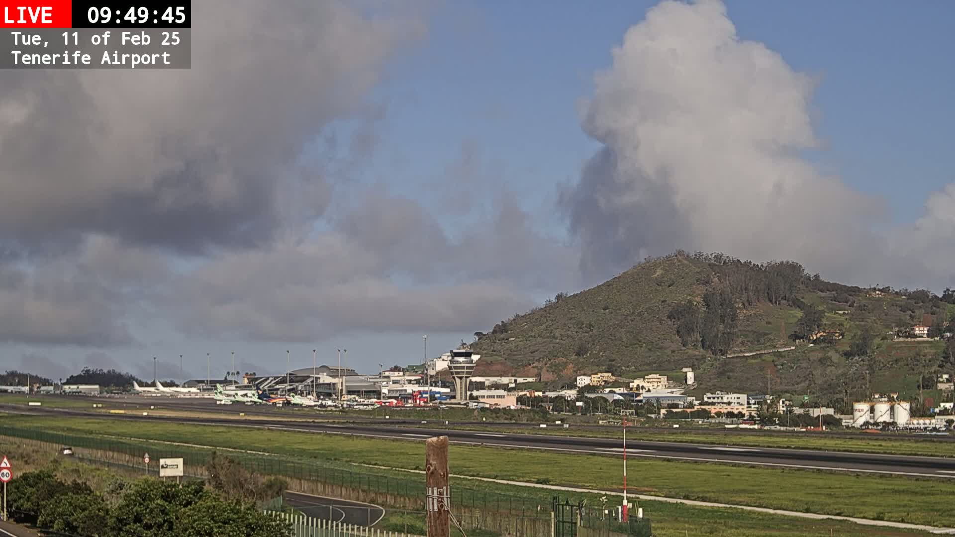 A partly cloudy day at an airport shows several airplanes parked on the tarmac, with a hill in the background.