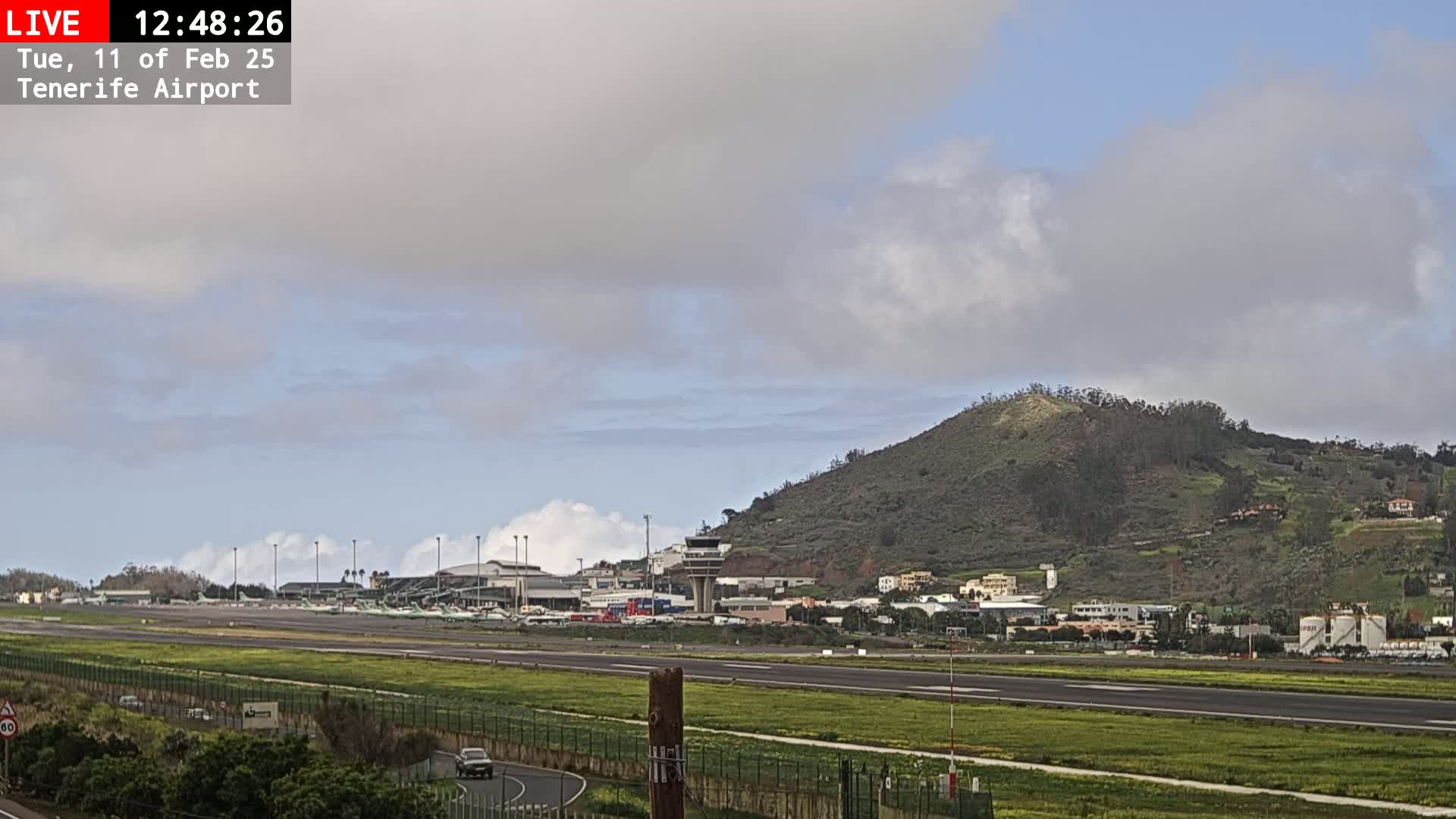 A mostly cloudy sky overlooks an airport runway with several planes parked, and a green hill in the background.