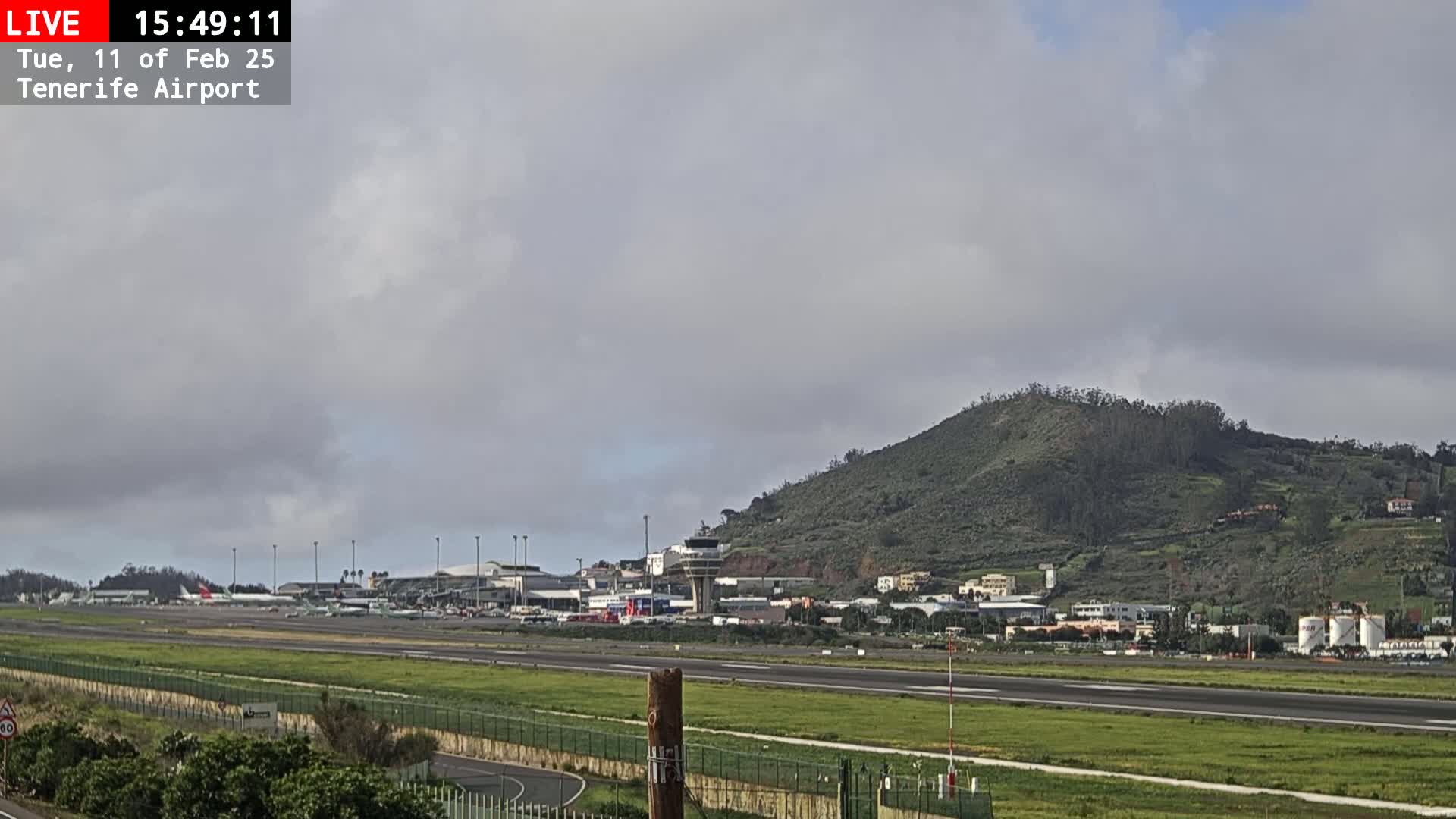 A long runway stretches towards an airport terminal and control tower, all situated at the base of a verdant hill under an overcast sky.