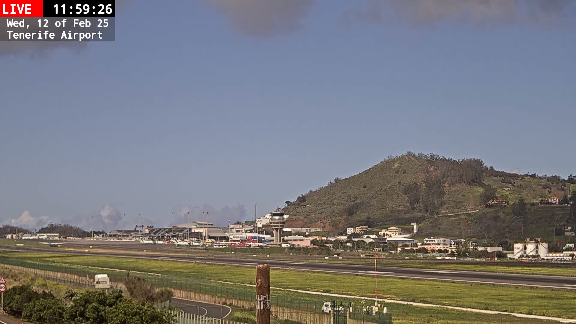 A long shot of an airport runway with several planes visible, under a mostly clear sky, with a large, green hill in the background.