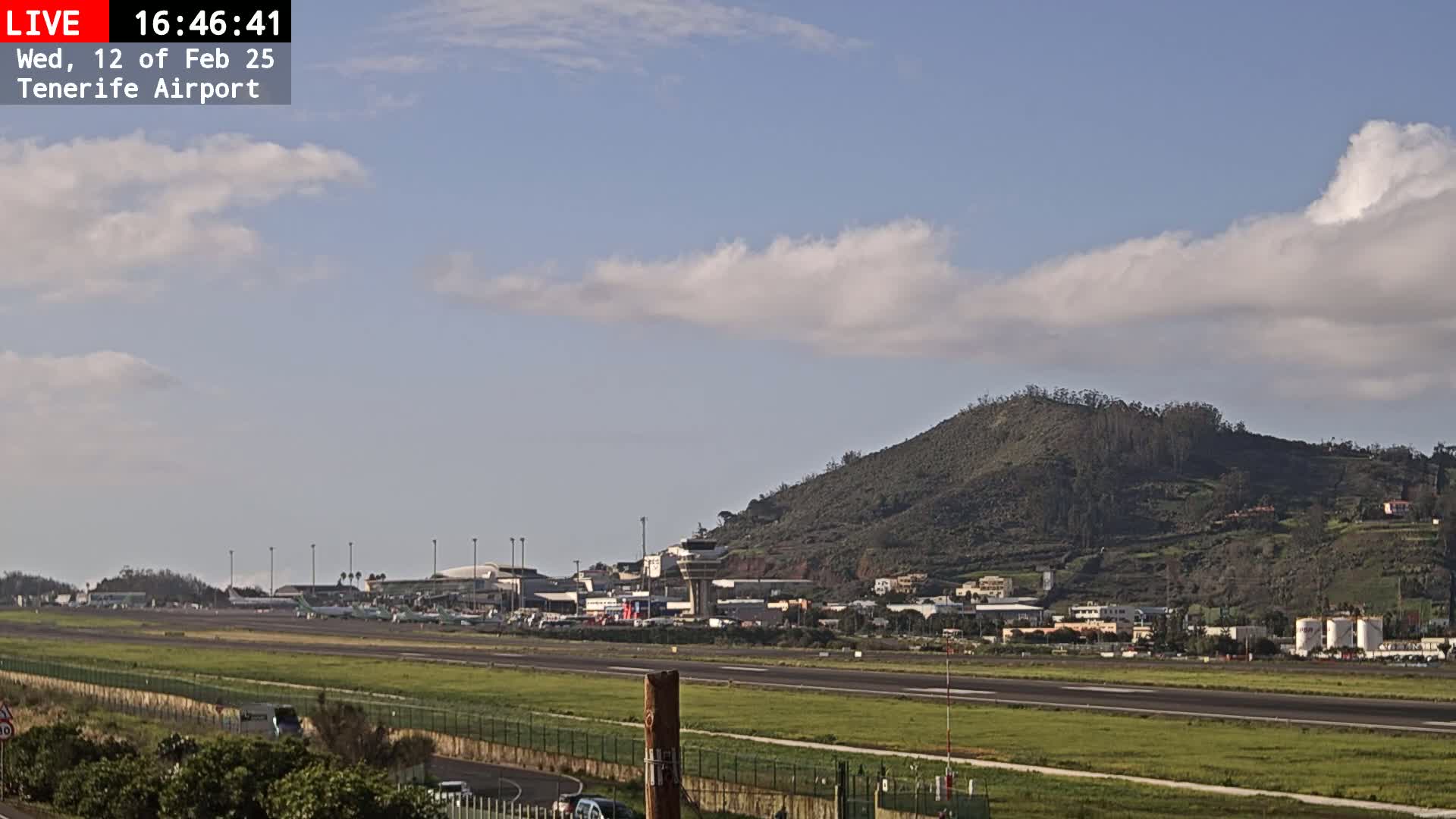 A mostly sunny day at an airport shows several airplanes on the tarmac with a large, green hill in the background.