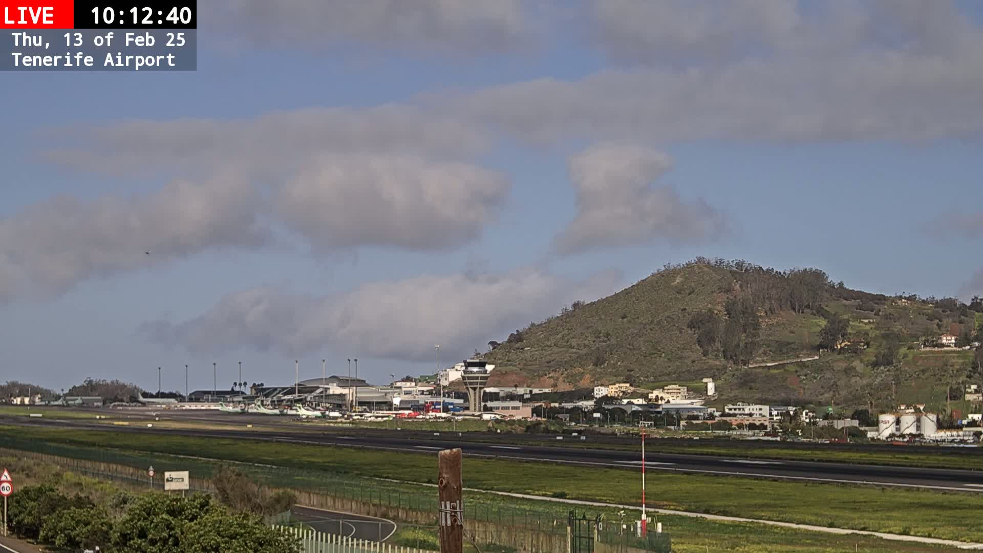 A partly cloudy day at an airport with a runway, terminal buildings, and a hill in the background.