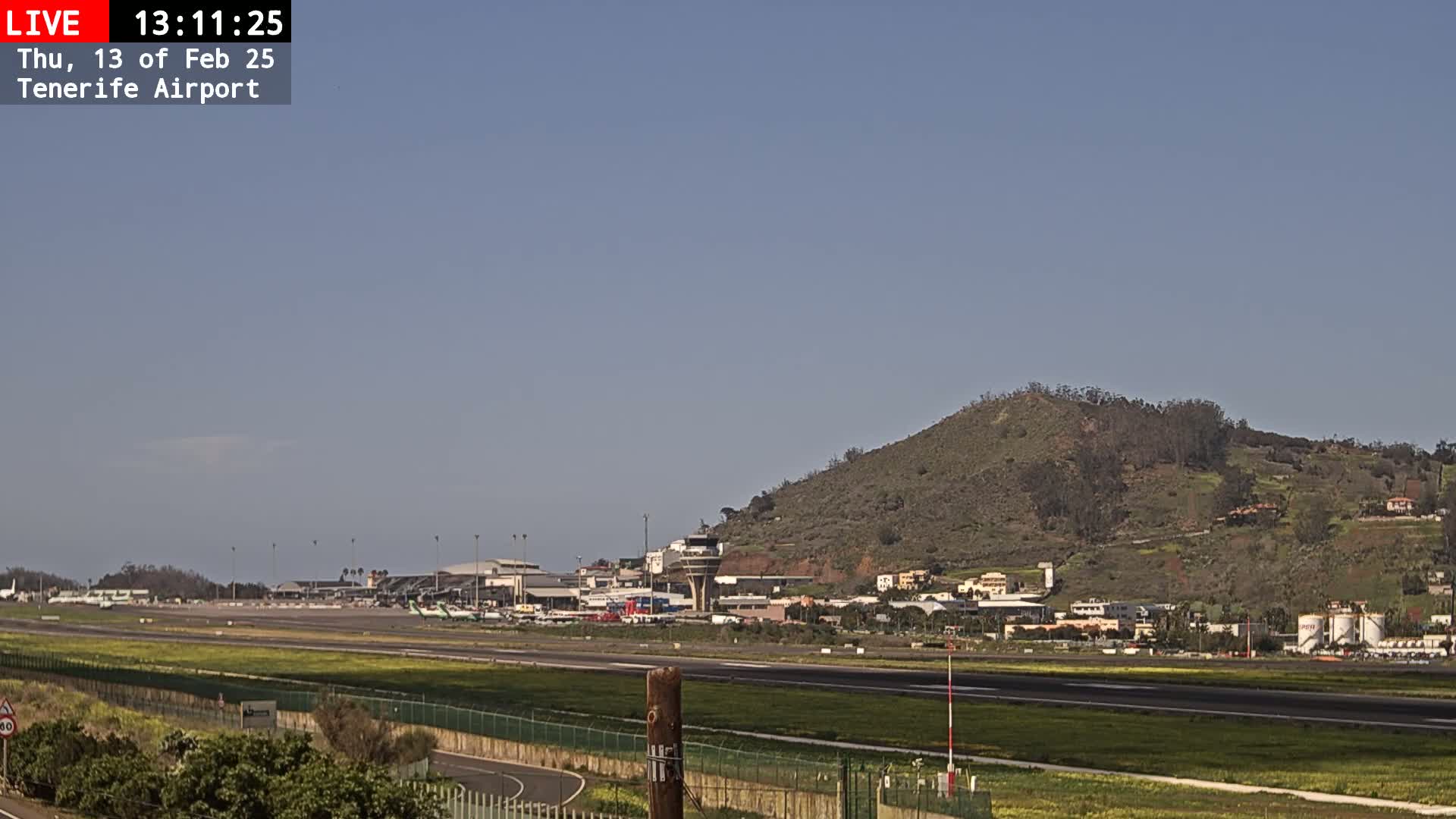 An airport runway and terminal are visible beneath a clear blue sky, with a large, sparsely vegetated hill in the background.