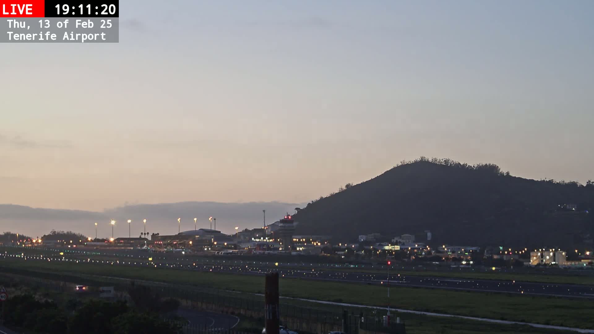 A dimly lit airport runway and terminal are visible beneath a dusky sky, with a dark hill rising in the background.