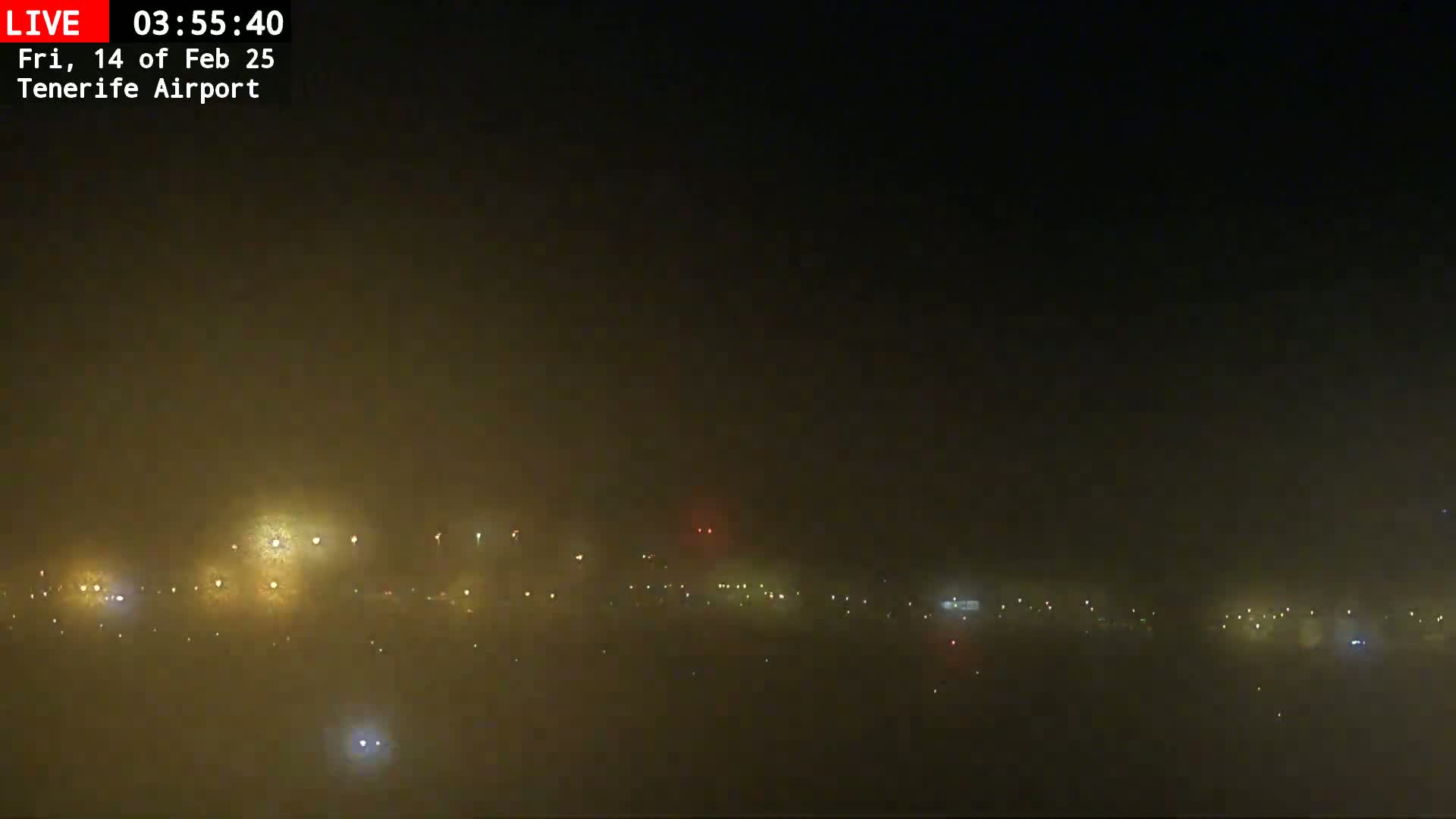 A nighttime view of an airport runway, obscured by heavy fog, with faint lights visible in the distance.