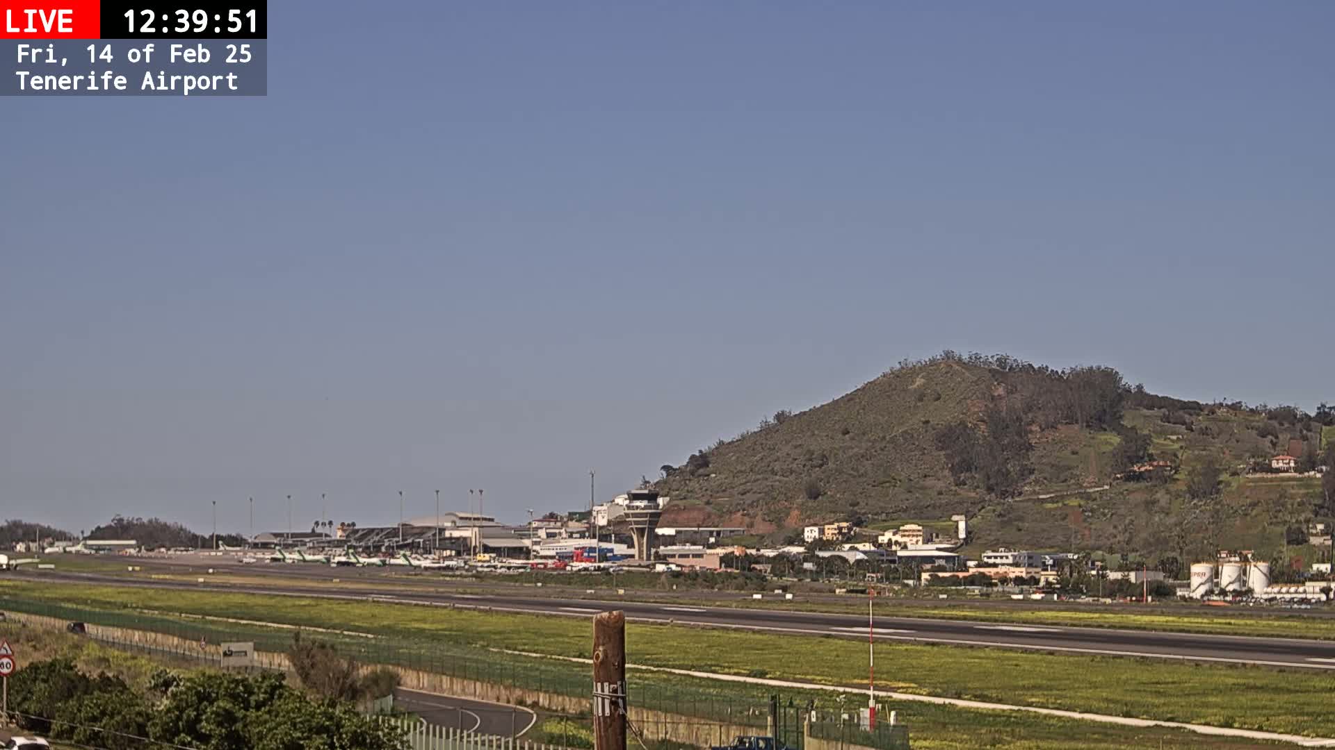 A sunny day at Tenerife Airport shows several airplanes parked on the tarmac with a runway in the foreground and a hill in the background.