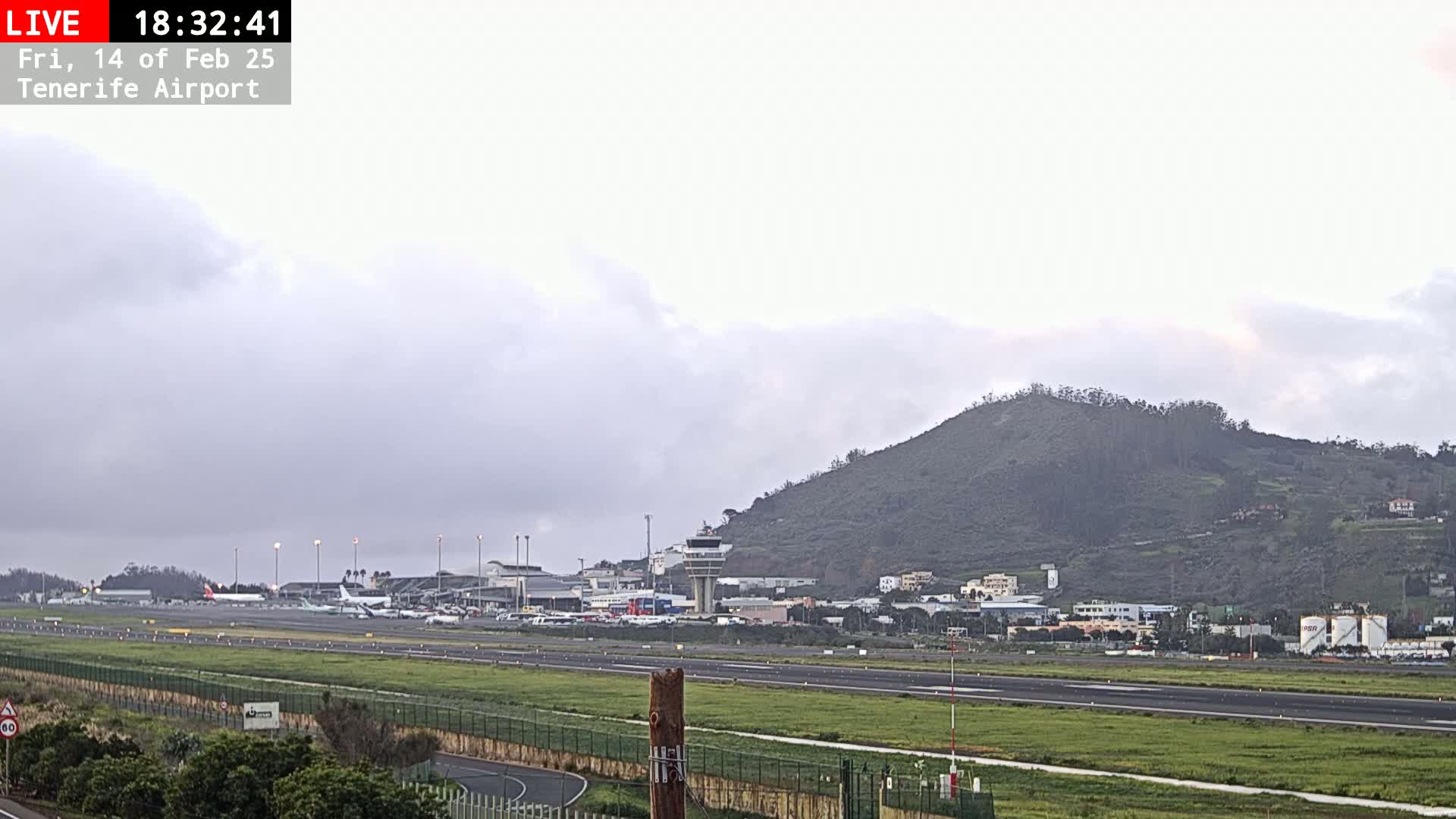 An overcast sky hangs over an airport with several airplanes on the tarmac, backed by a low, dark green hill.