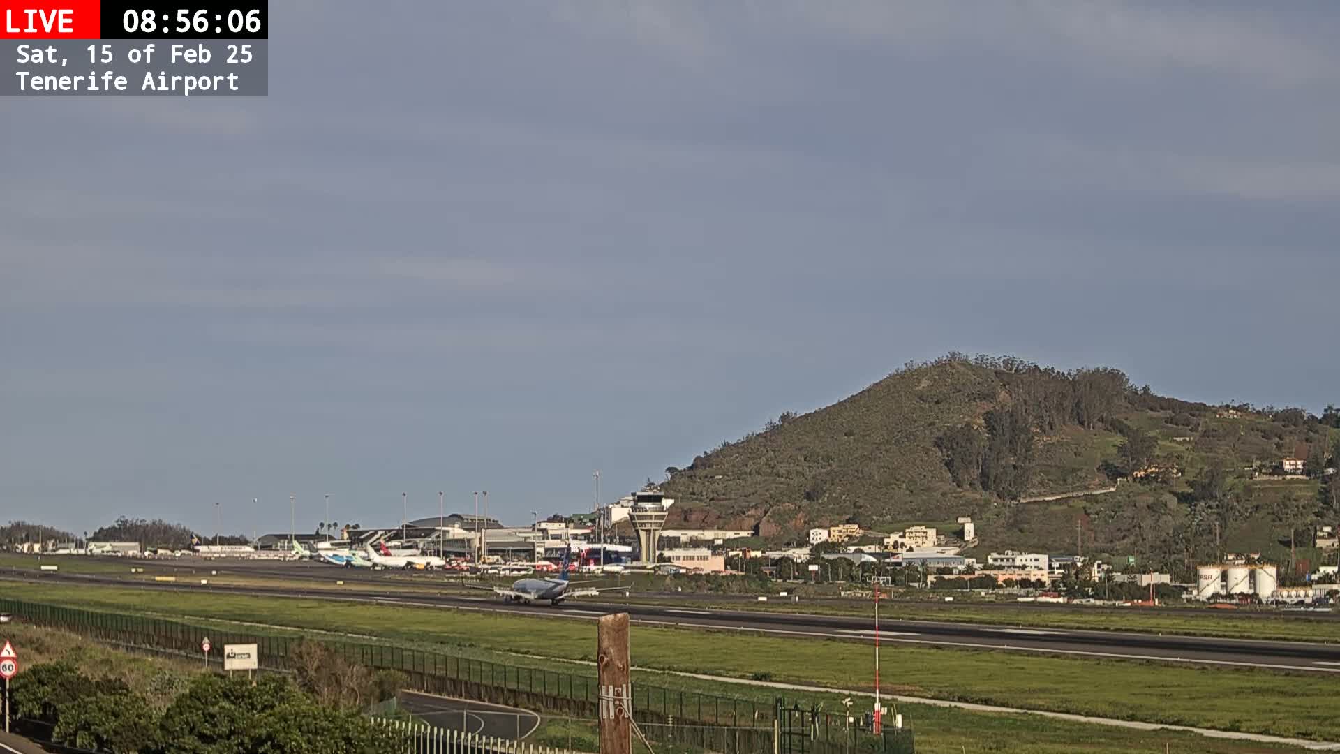 An airport runway with several airplanes, a control tower, and surrounding buildings is visible under a mostly clear sky.