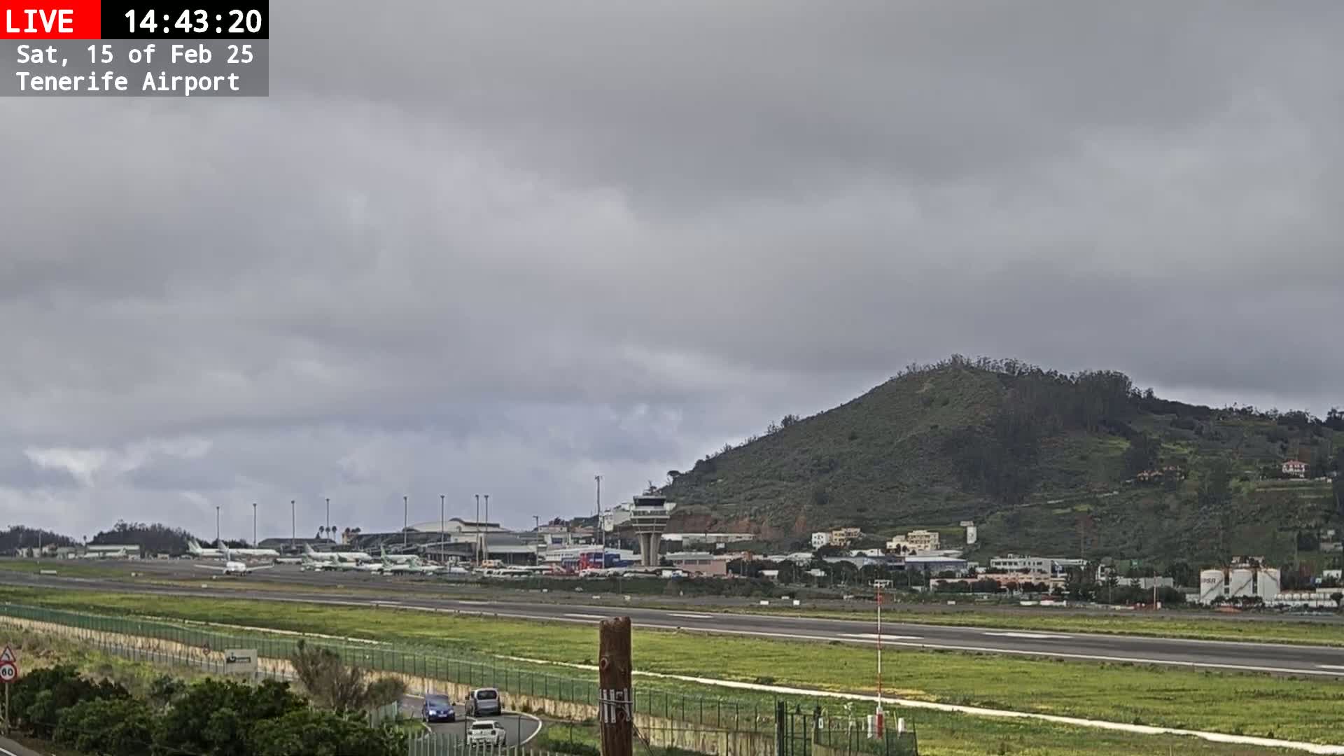 A view of an airport runway with several airplanes parked and a hill in the background under an overcast sky.